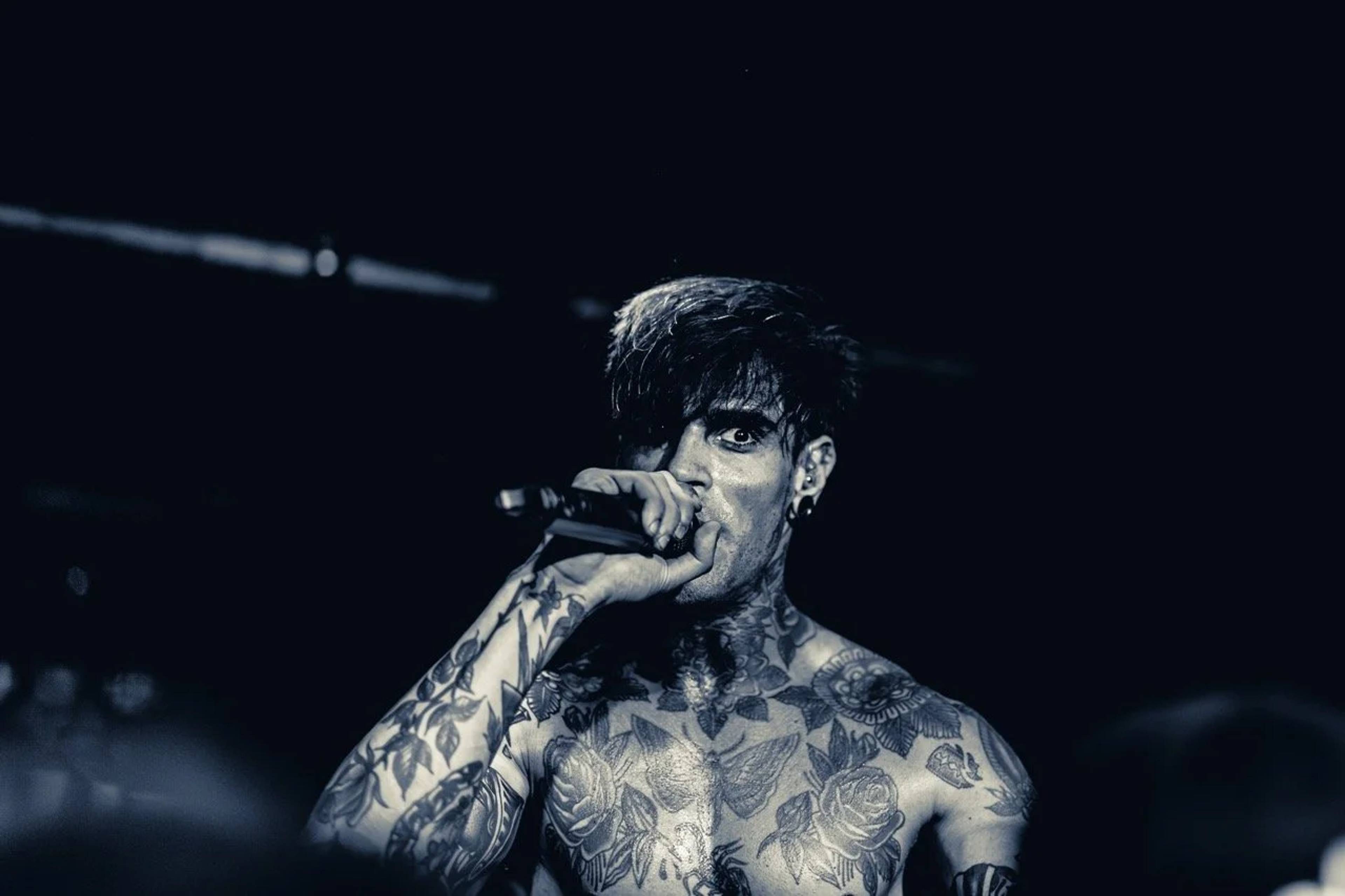 A shirtless tattooed vocalist performs on stage, holding a microphone with intensity under dramatic blue-tinted stage lighting.