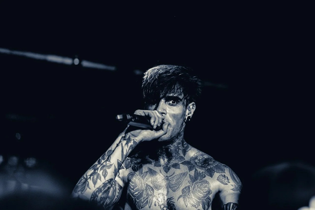 A shirtless tattooed vocalist performs on stage, holding a microphone with intensity under dramatic blue-tinted stage lighting.