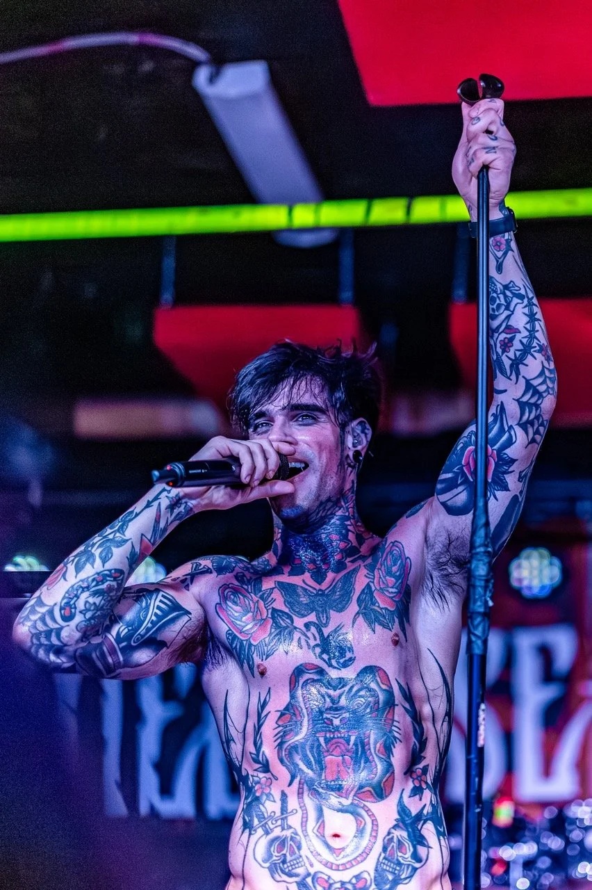 A shirtless tattooed singer performs on stage at Empire Live Underground, holding a microphone with his fist raised, illuminated by red and green stage lighting.