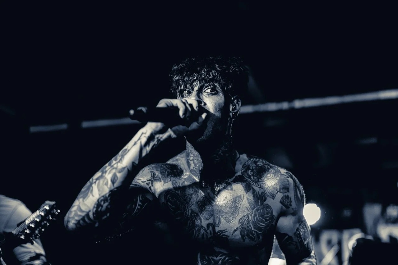 A tattooed male vocalist performing on stage with a microphone, lit in monochrome against a dark background with stage lights.