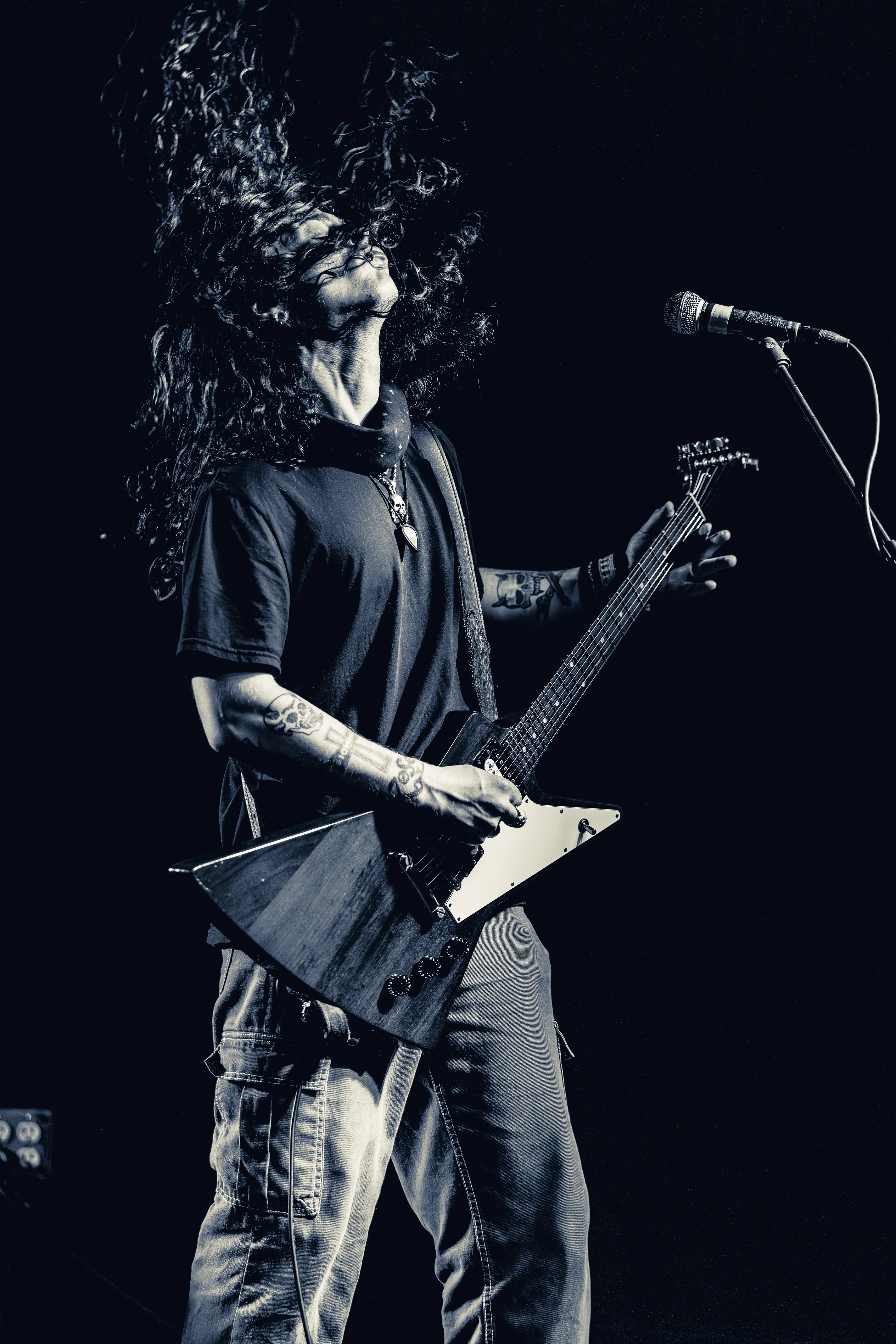 A long-haired musician on stage playing an angular electric guitar with microphone stand visible, lit against a dark background.