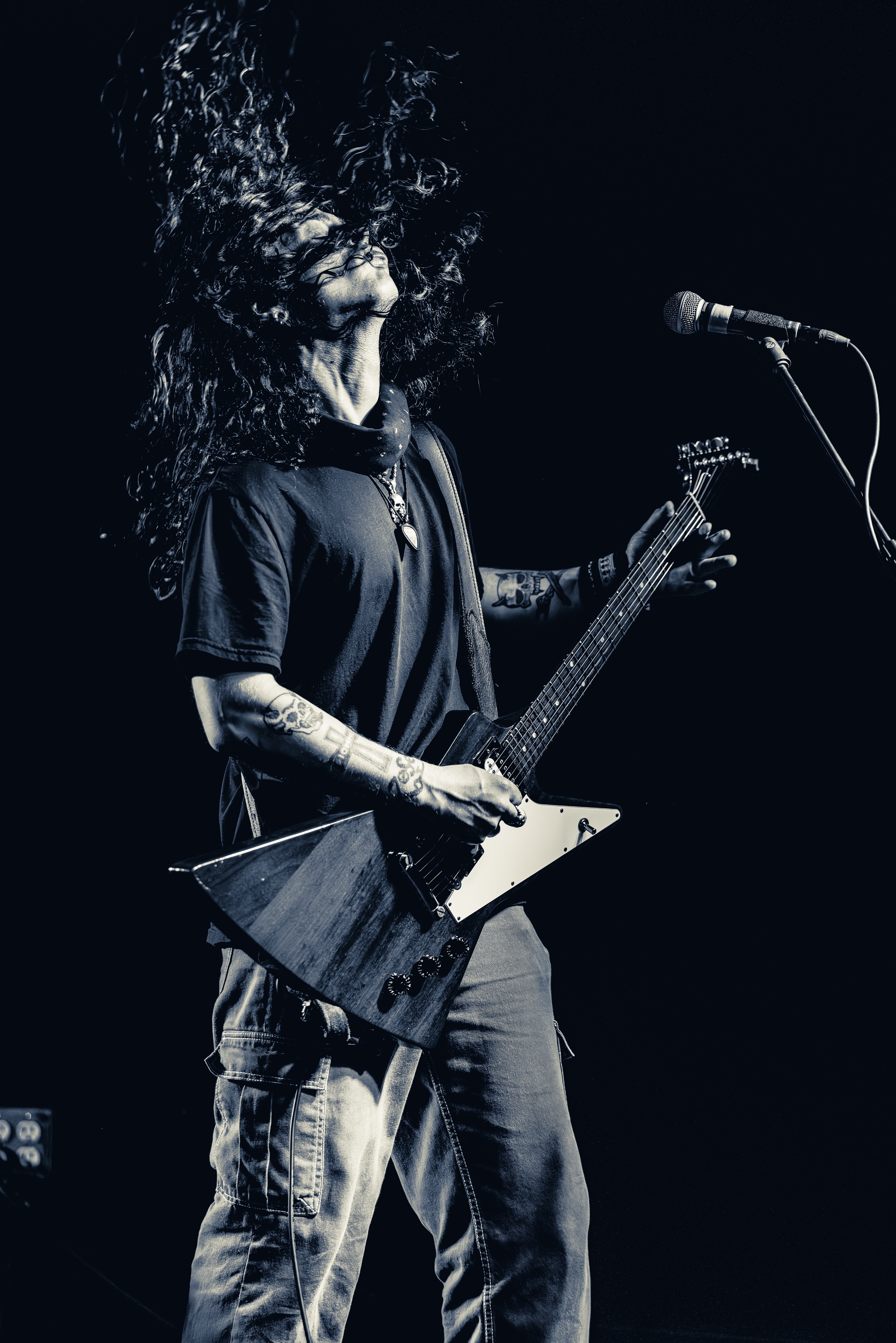A long-haired musician on stage playing an angular electric guitar with microphone stand visible, lit against a dark background.