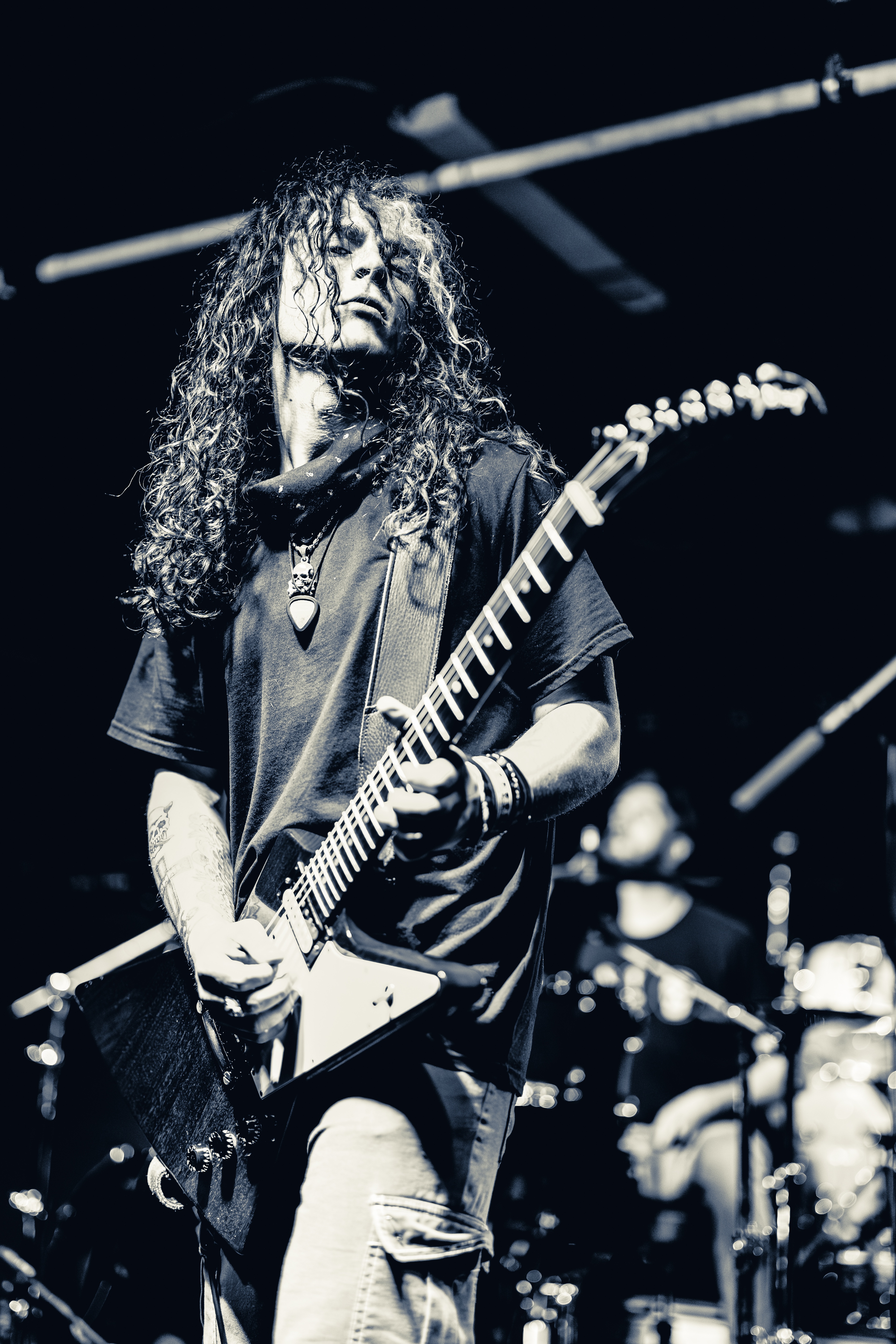 A long-haired guitarist in a dark t-shirt plays an electric guitar on stage, illuminated by white stage lighting against a dark background with drum kit visible behind.