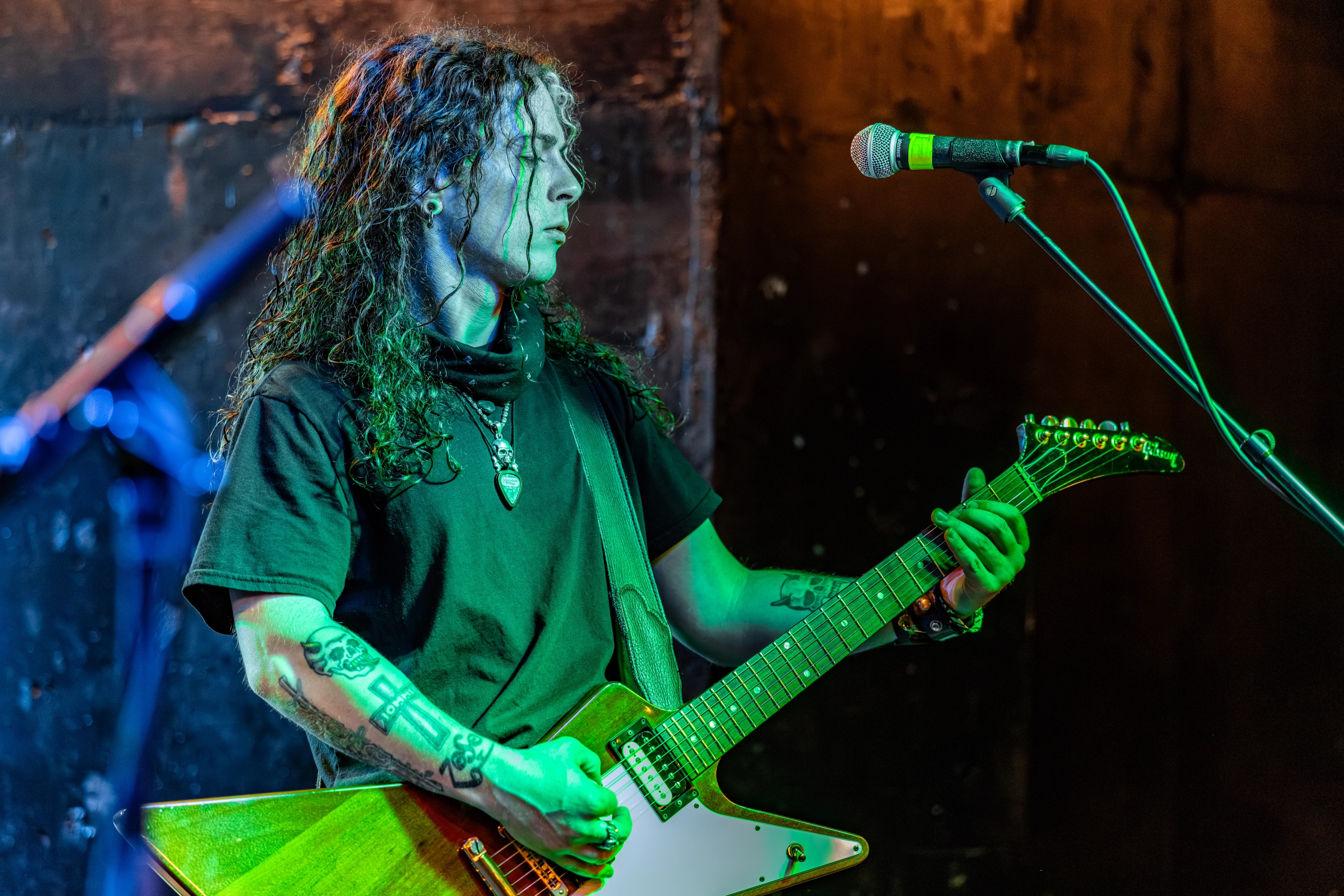 A long-haired guitarist on stage holding a guitar, lit in green and blue stage lights, eyes focused downward while playing.