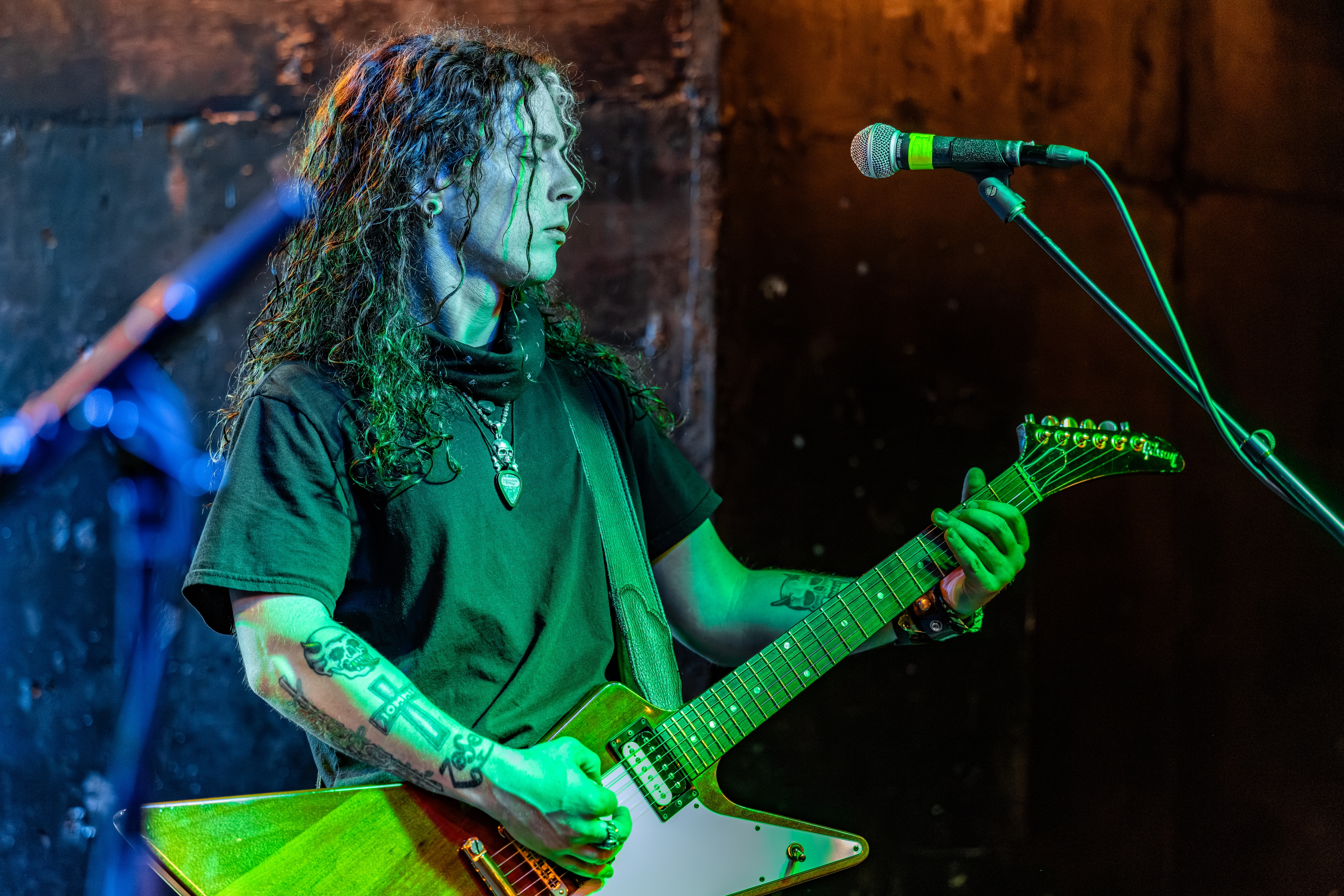 A long-haired guitarist on stage holding a guitar, lit in green and blue stage lights, eyes focused downward while playing.