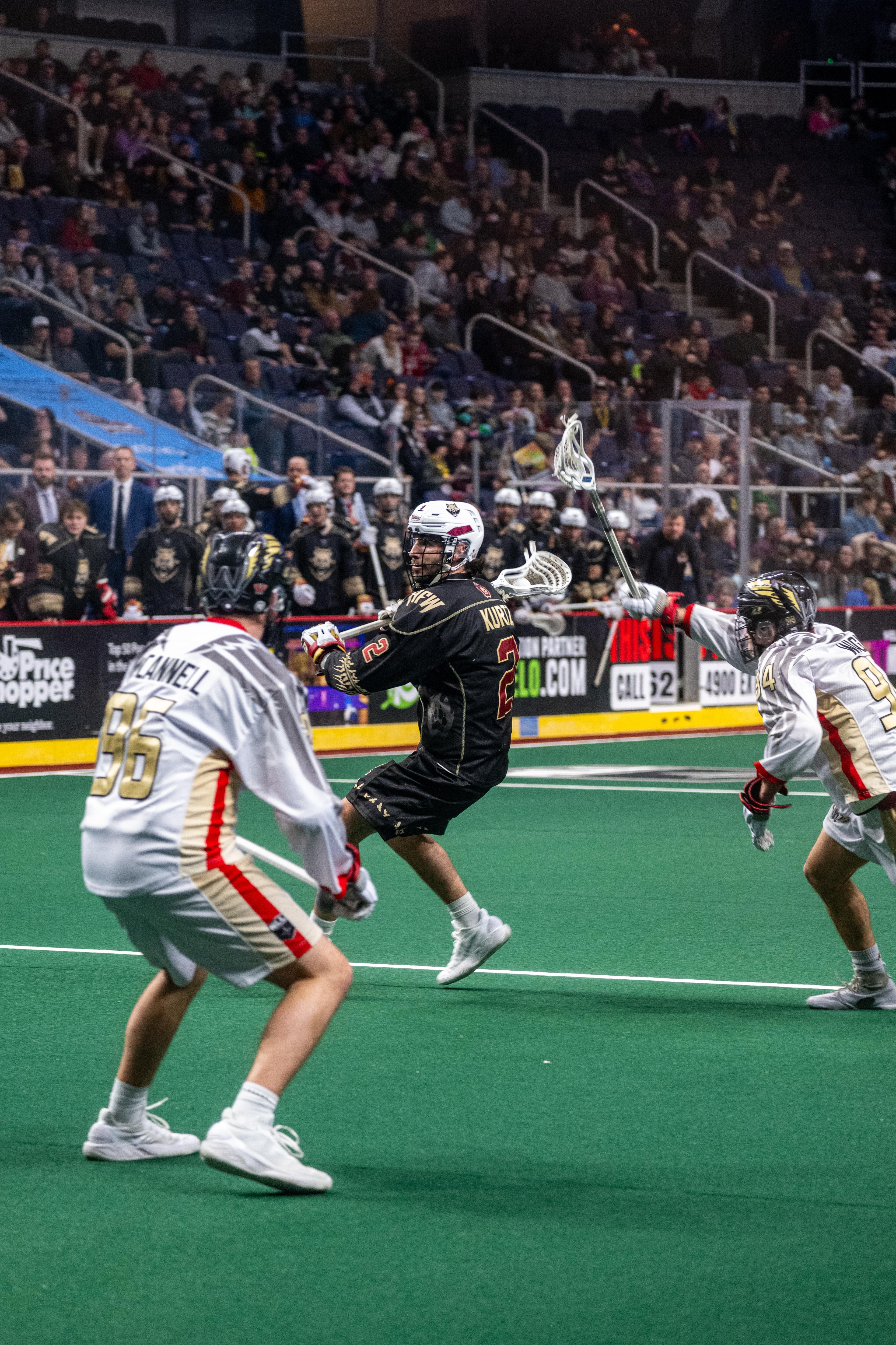 A lacrosse player in black jersey with stick raised evades two white-jersey defenders on indoor field with crowd in stadium background.