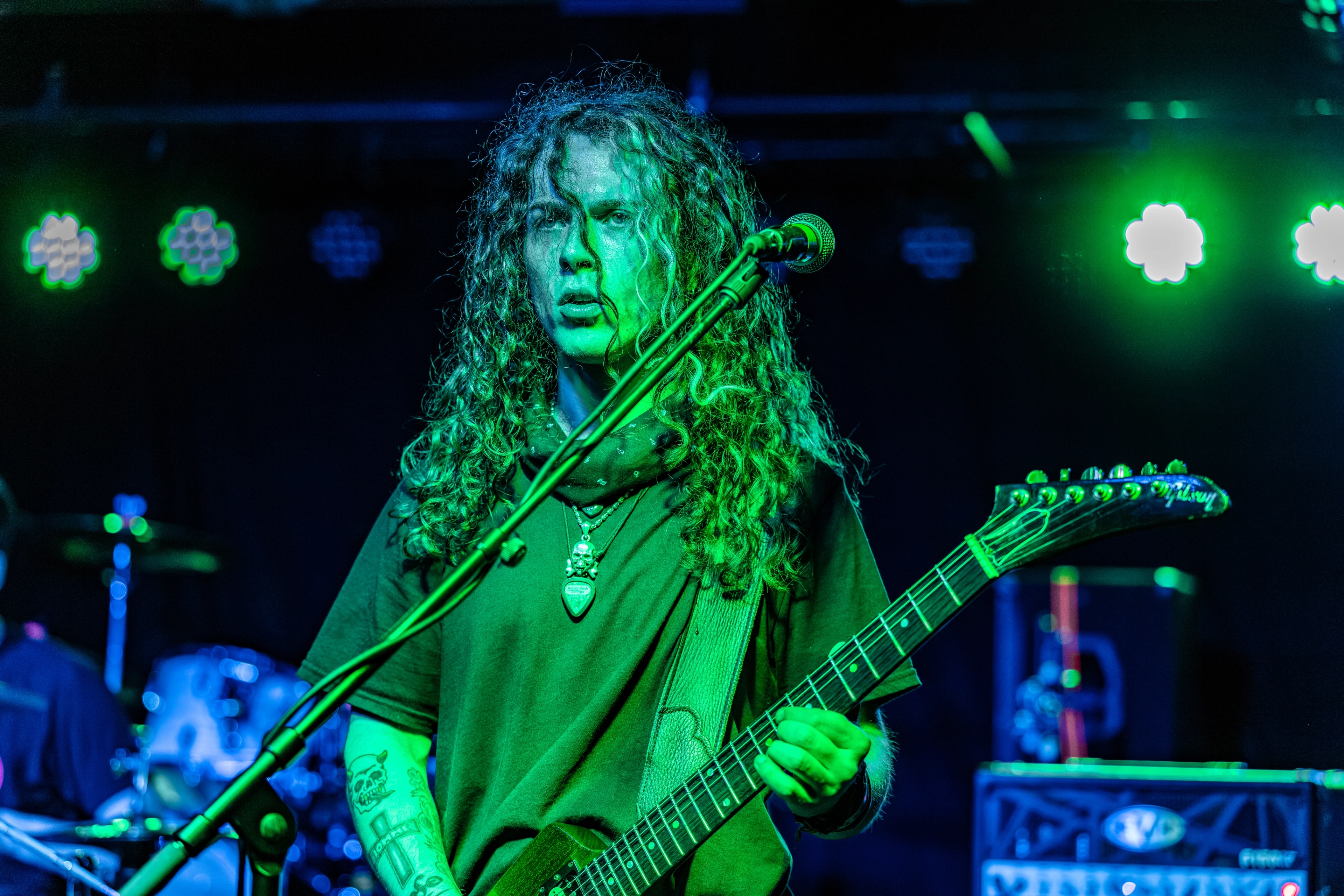 A long-haired guitarist performing on stage under green and blue stage lighting, holding an electric guitar with focused expression.