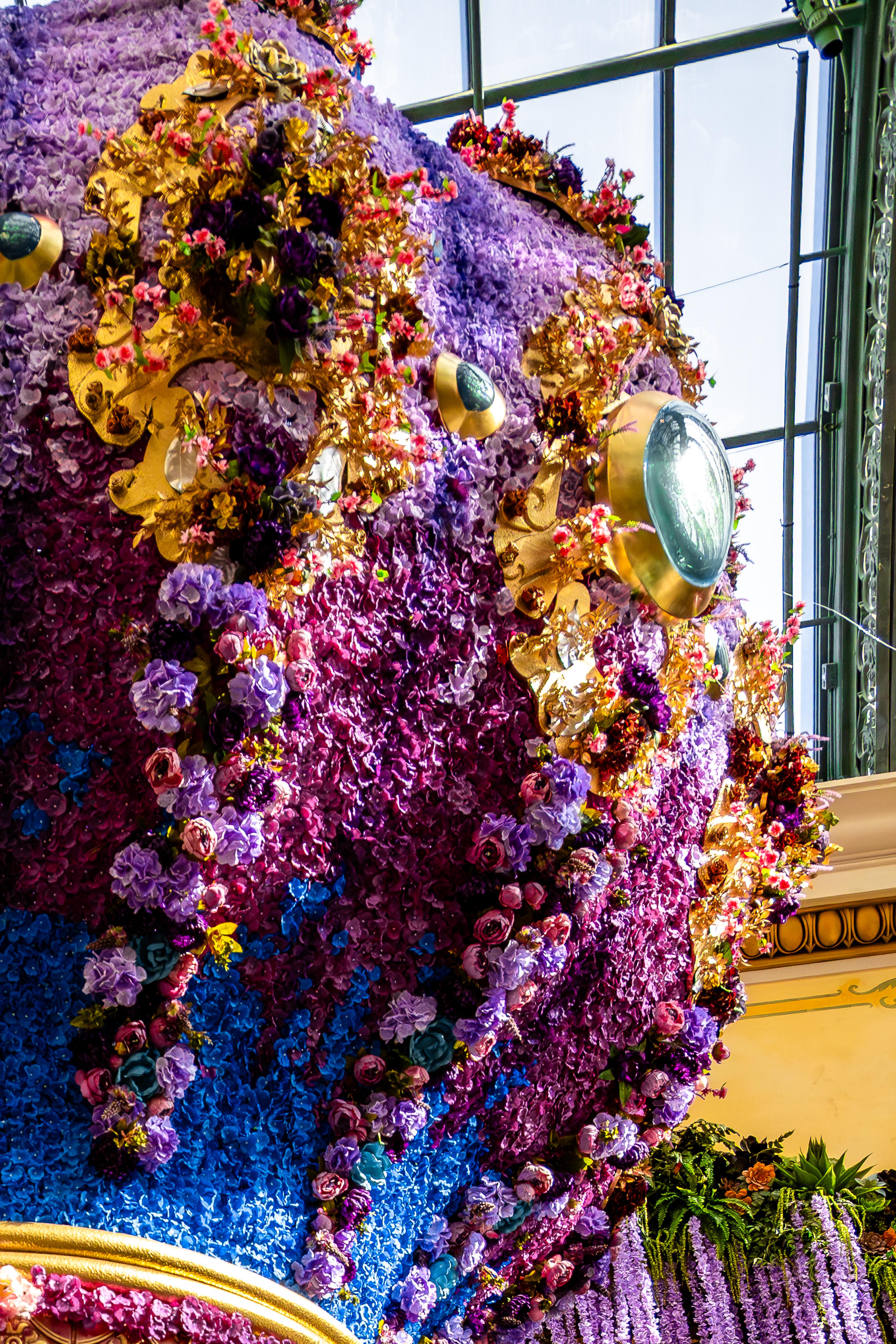 A massive floral wall installation with purple, pink, and blue flowers interspersed with gold ornamental elements and brass mirrors, lit by natural light from large windows.