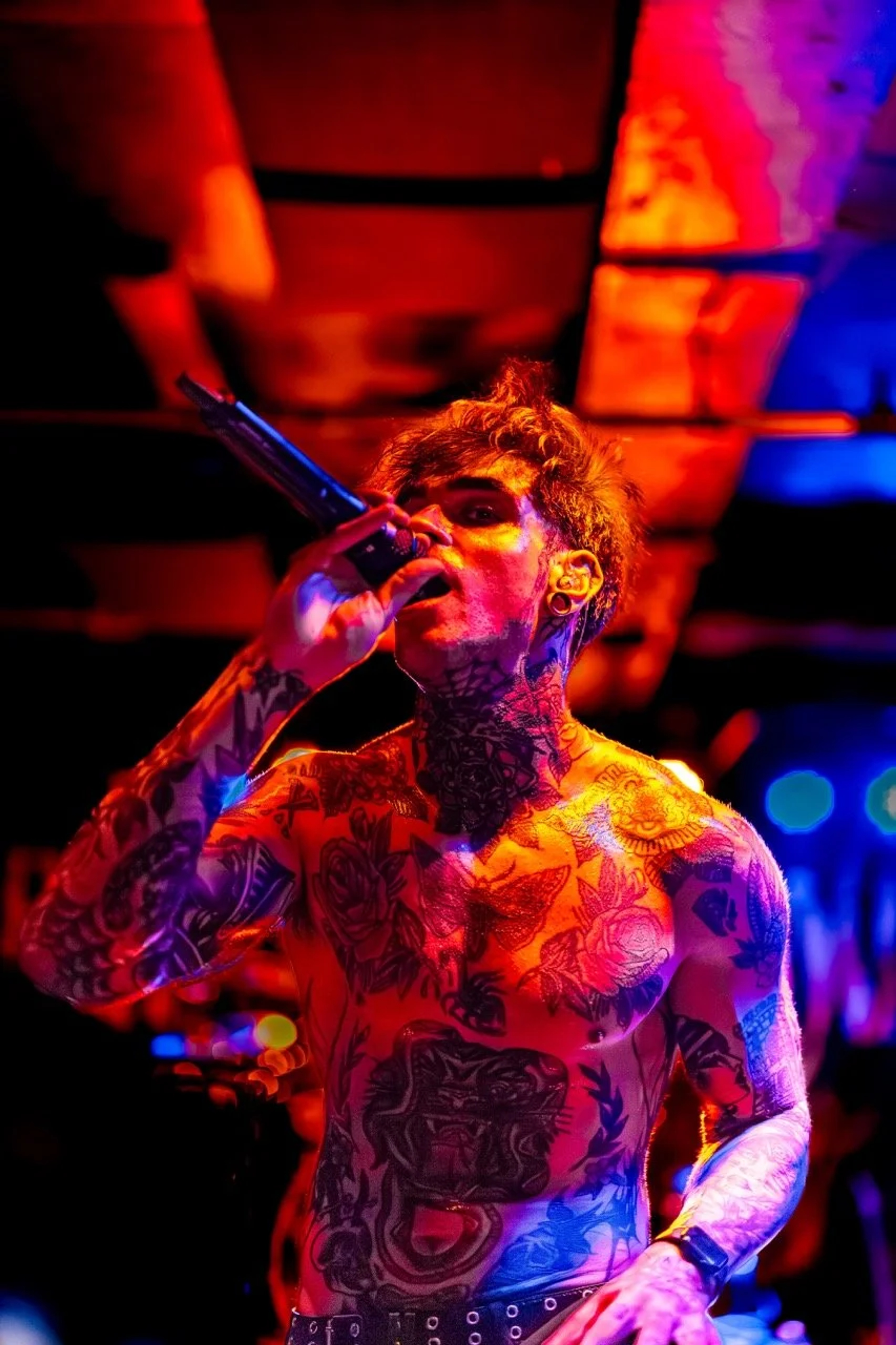 A shirtless tattooed singer performs on stage under intense red, orange, and blue stage lighting, holding a microphone with head tilted back.