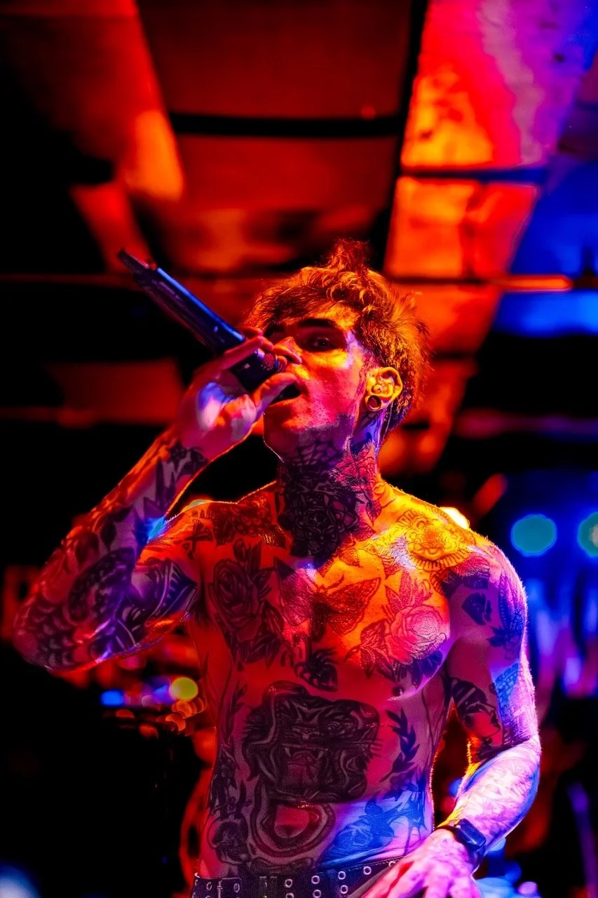 A shirtless tattooed singer performs on stage under intense red, orange, and blue stage lighting, holding a microphone with head tilted back.