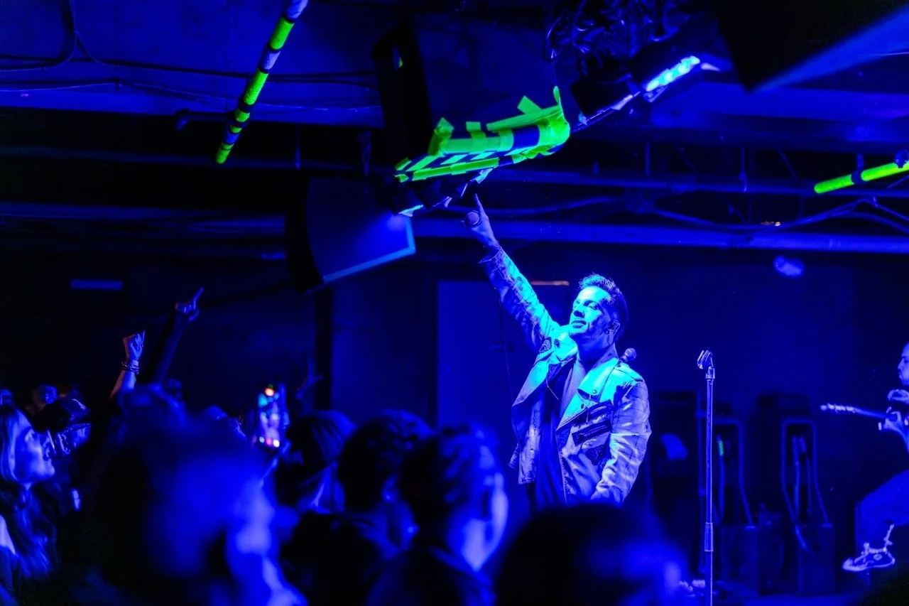 A musician on stage with arm raised, holding a guitar overhead under blue and neon green stage lighting, surrounded by a crowd in an underground venue.