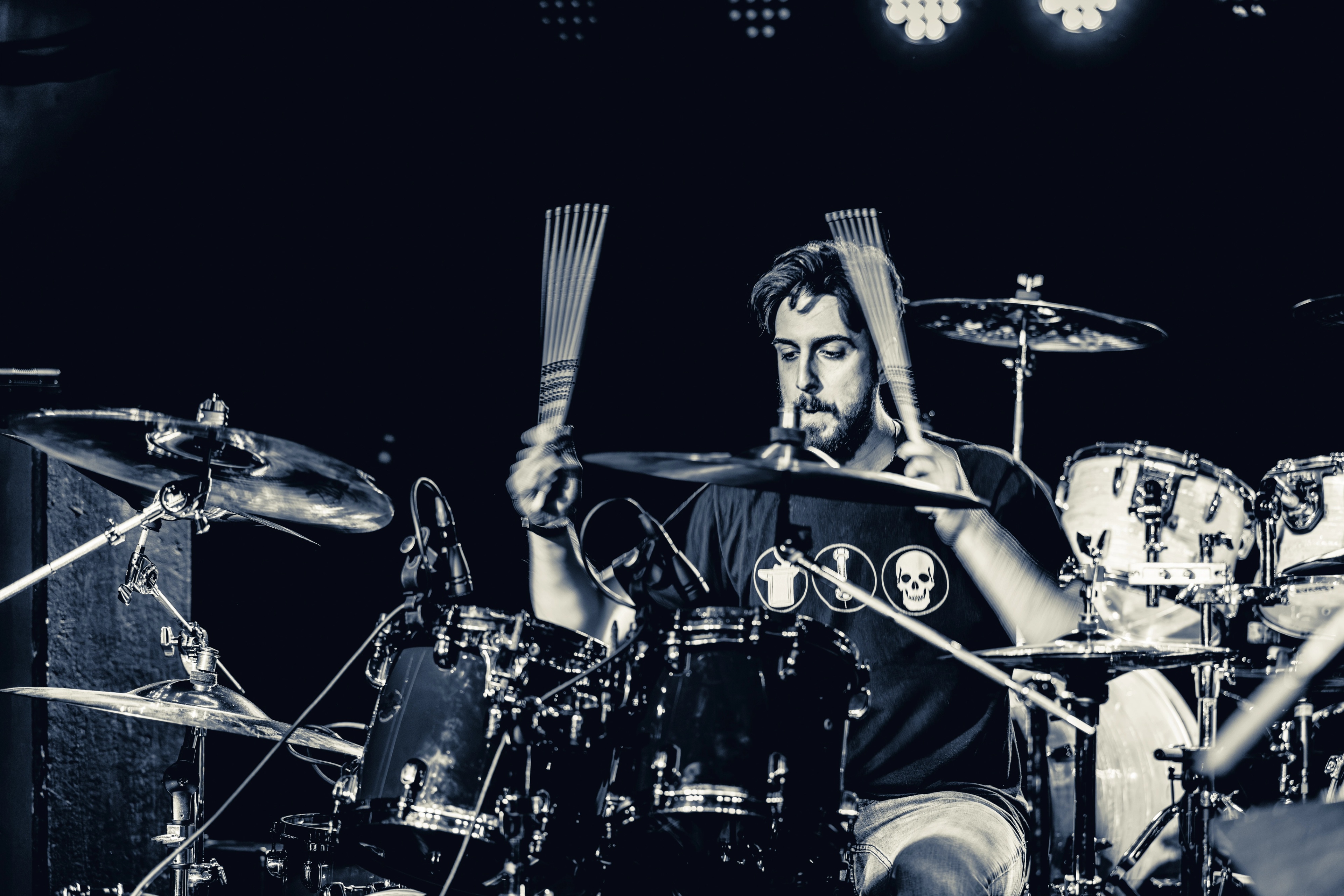 A bearded drummer in a black graphic tee plays cymbals and drums on stage under bright white stage lights during a live performance.