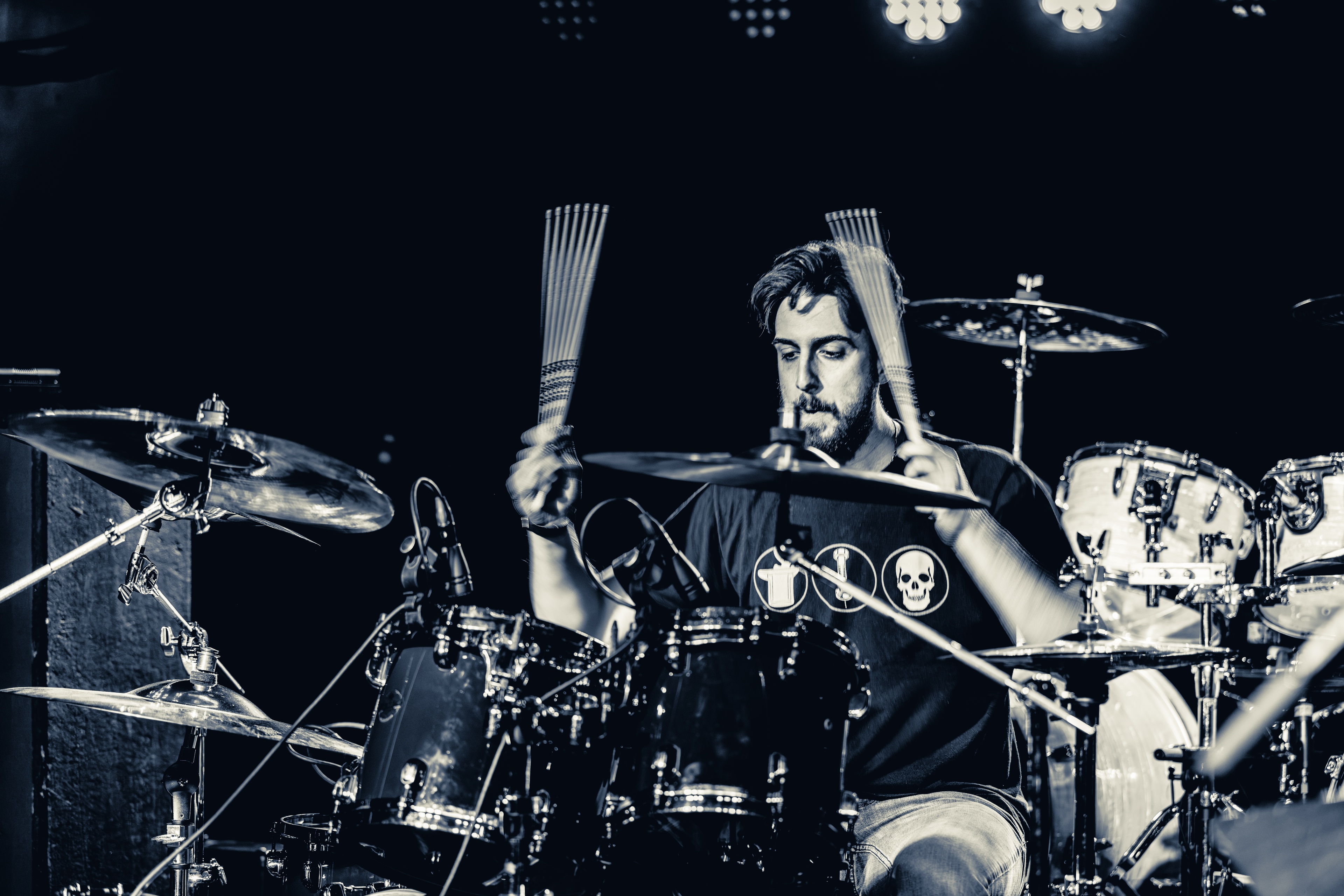 A bearded drummer in a black graphic tee plays cymbals and drums on stage under bright white stage lights during a live performance.