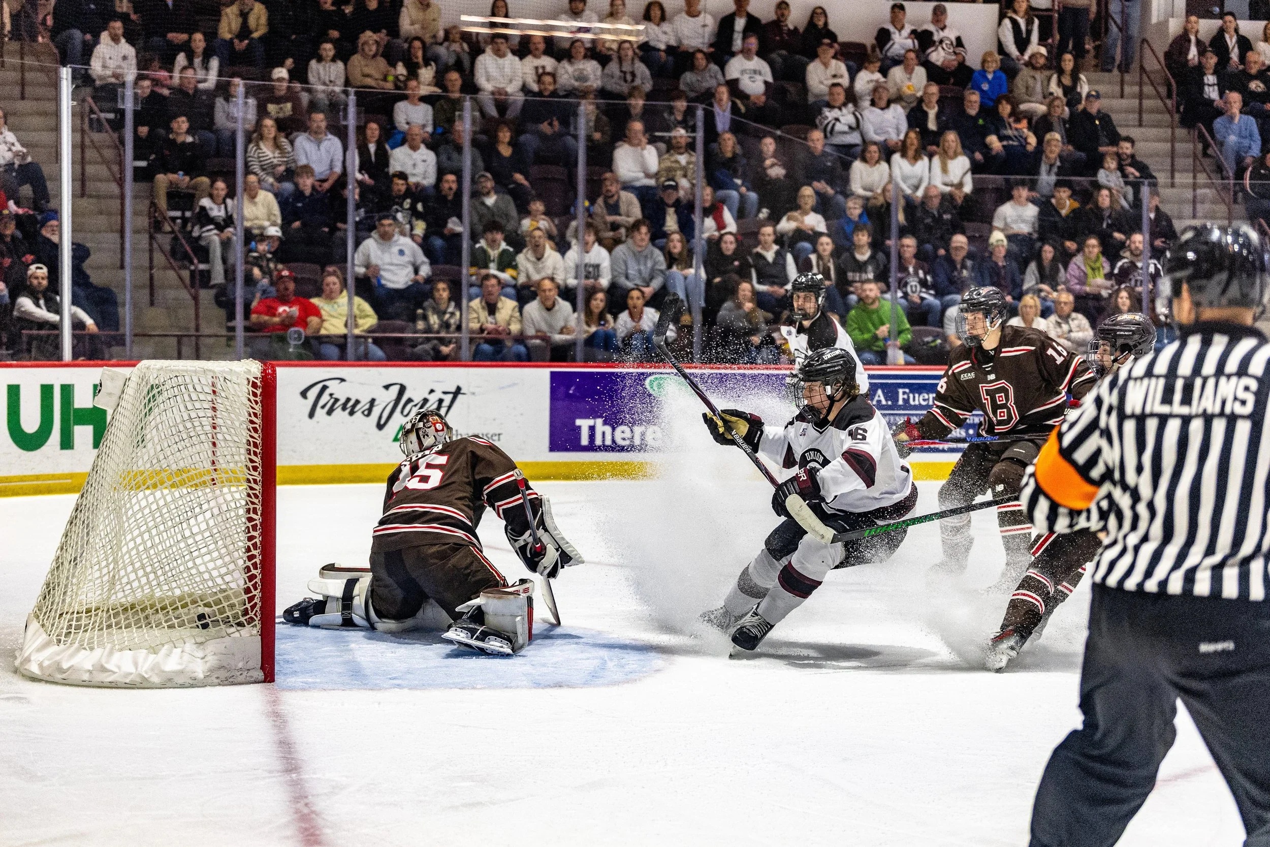 A hockey player in white jersey drives toward the goal while a goaltender in dark jersey crouches in the net, with ice spray and crowd visible in background.