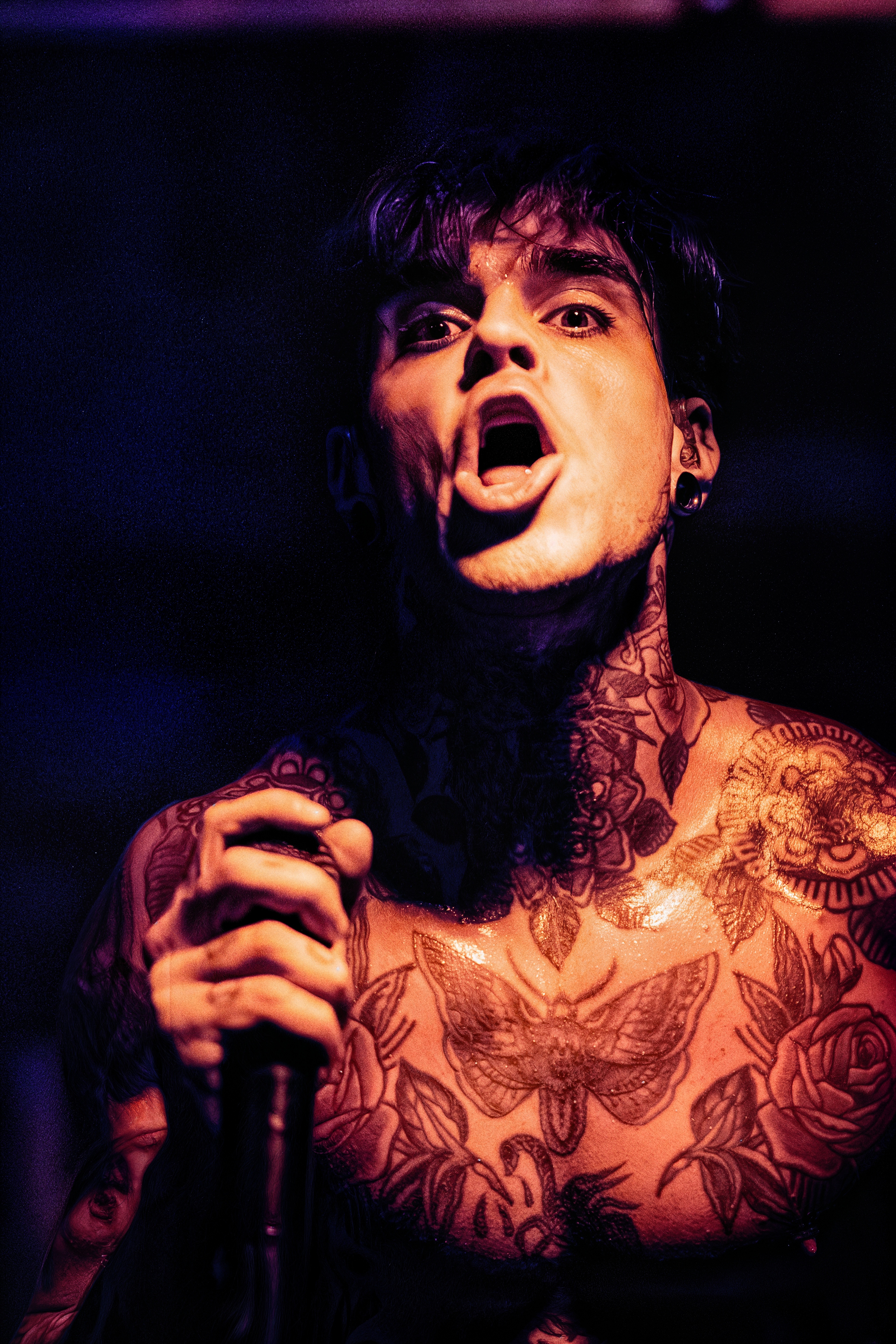 A shirtless vocalist with extensive tattoos grips a microphone, mouth open mid-performance, lit in warm orange and purple stage lighting against a dark background.