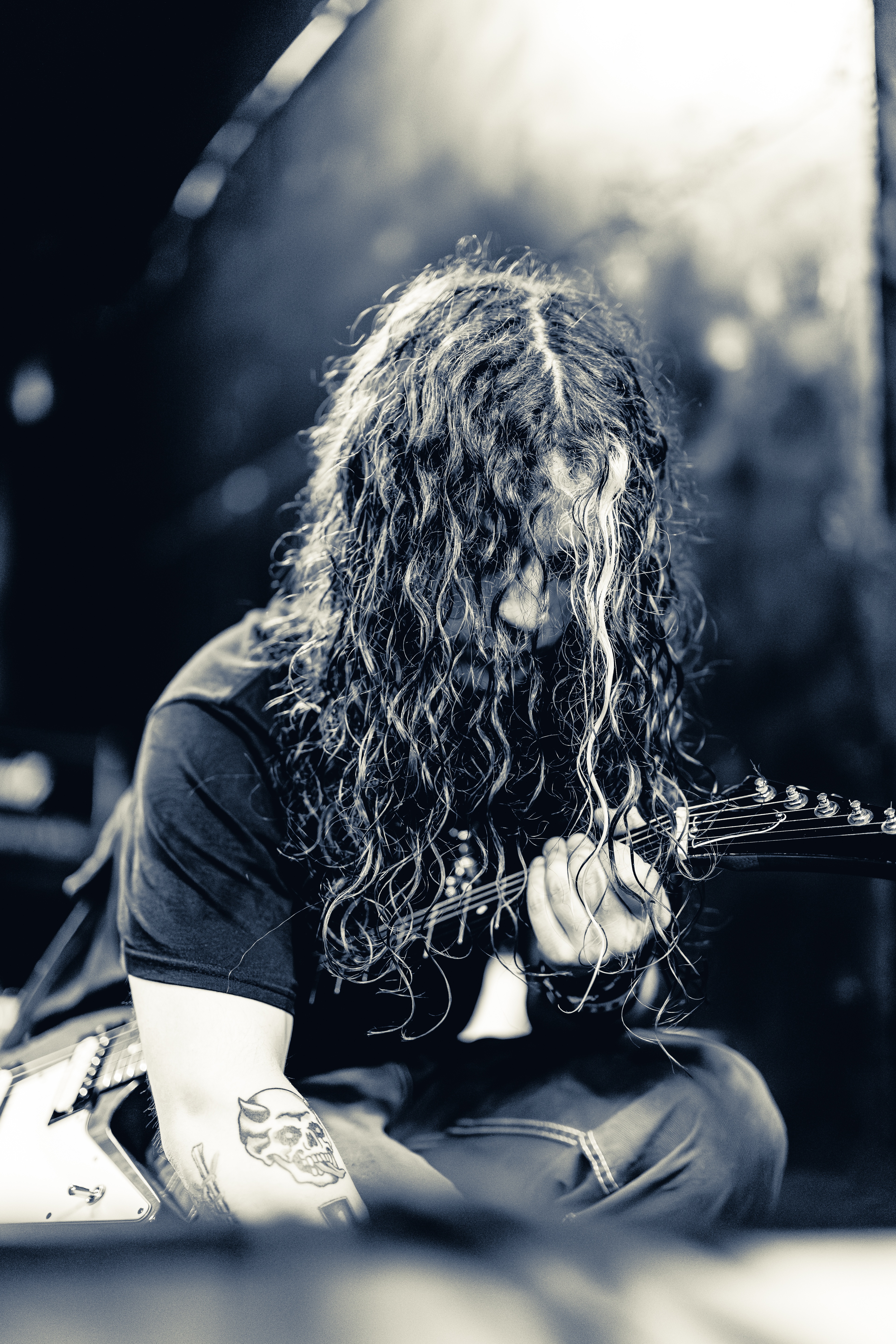 A long-haired guitarist performs on stage, leaning into an electric guitar with sweat-soaked hair backlit by stage lighting.
