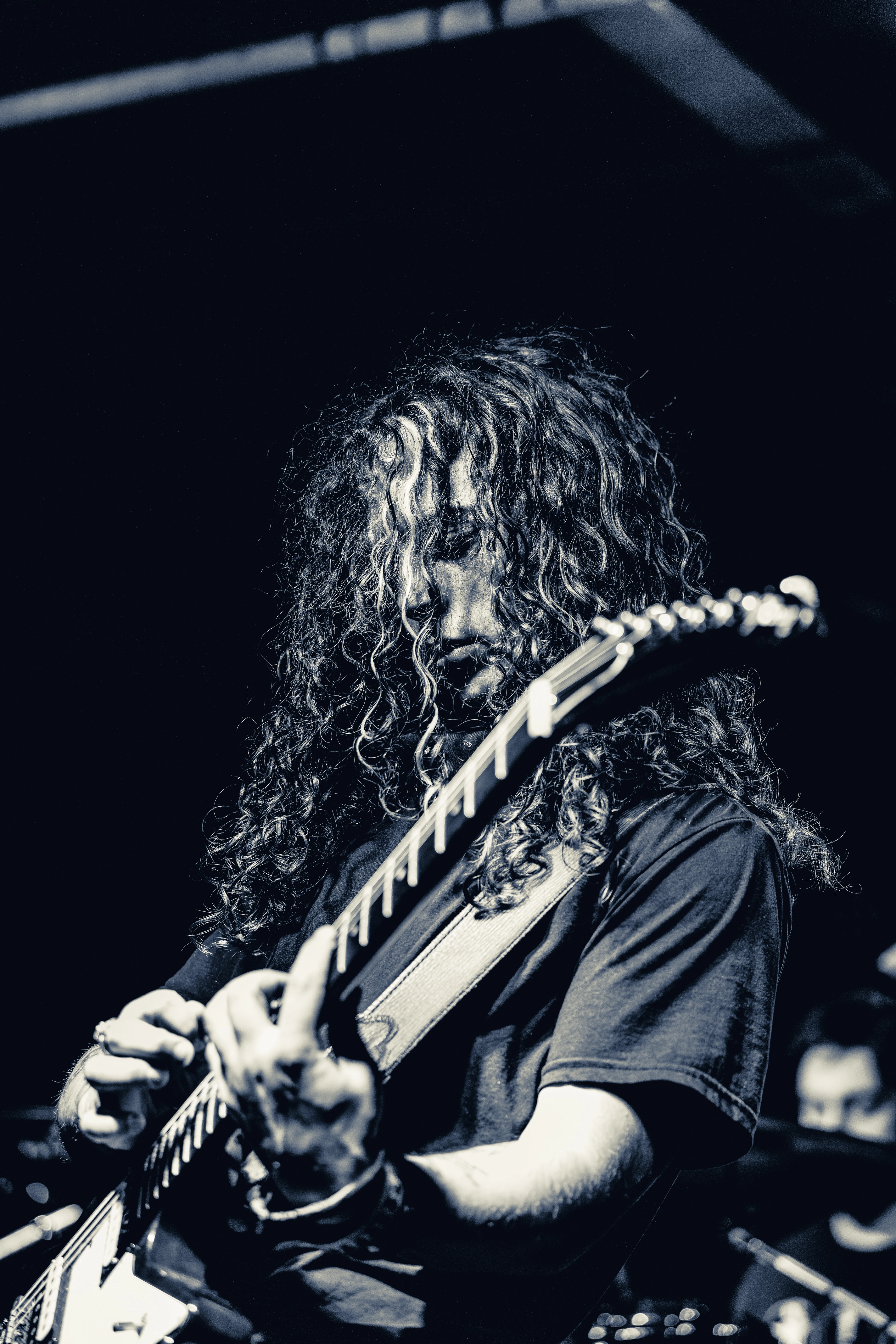 A long-haired guitarist performs on stage, focused downward while playing an electric guitar under dramatic stage lighting.