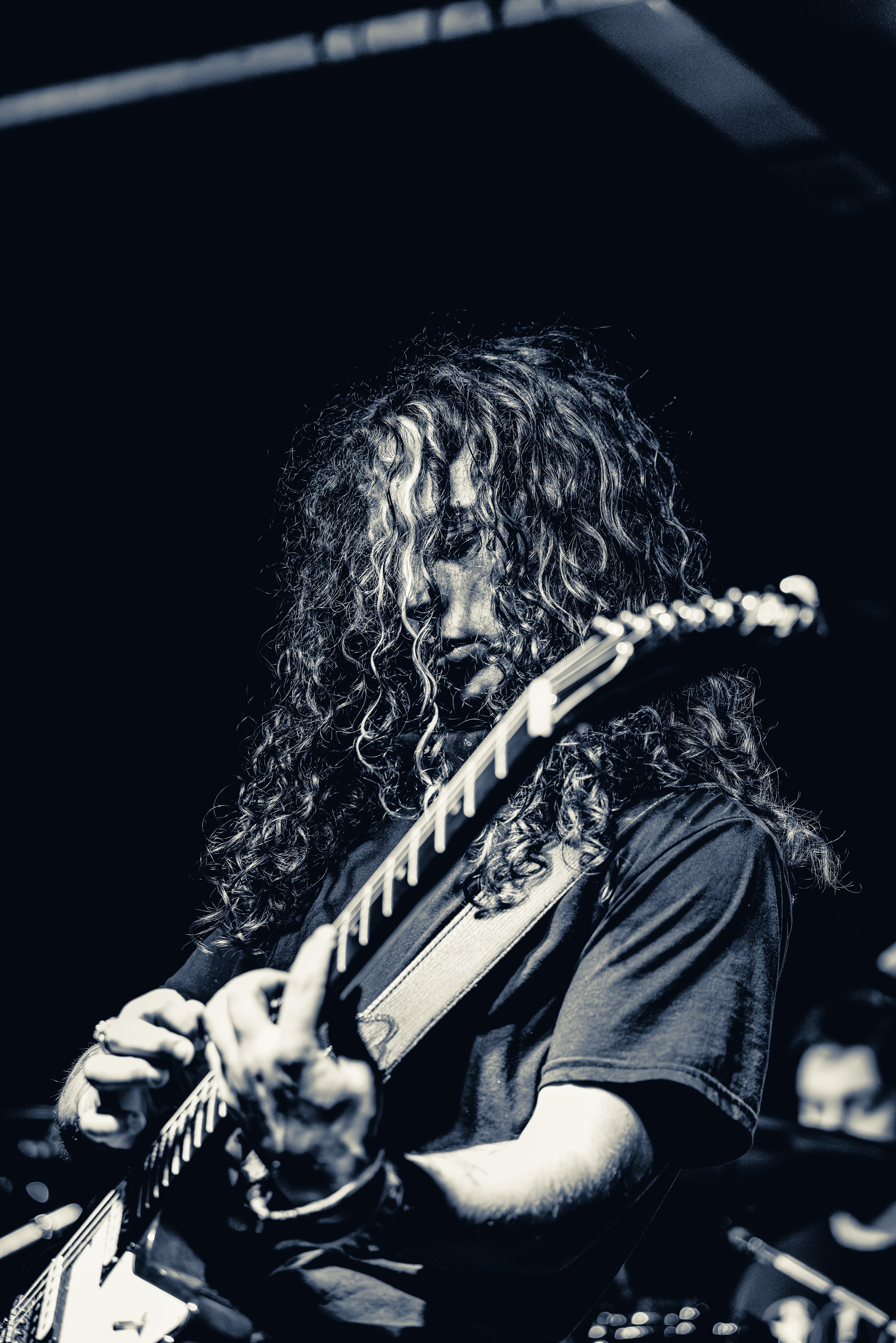 A long-haired guitarist performs on stage, focused downward while playing an electric guitar under dramatic stage lighting.