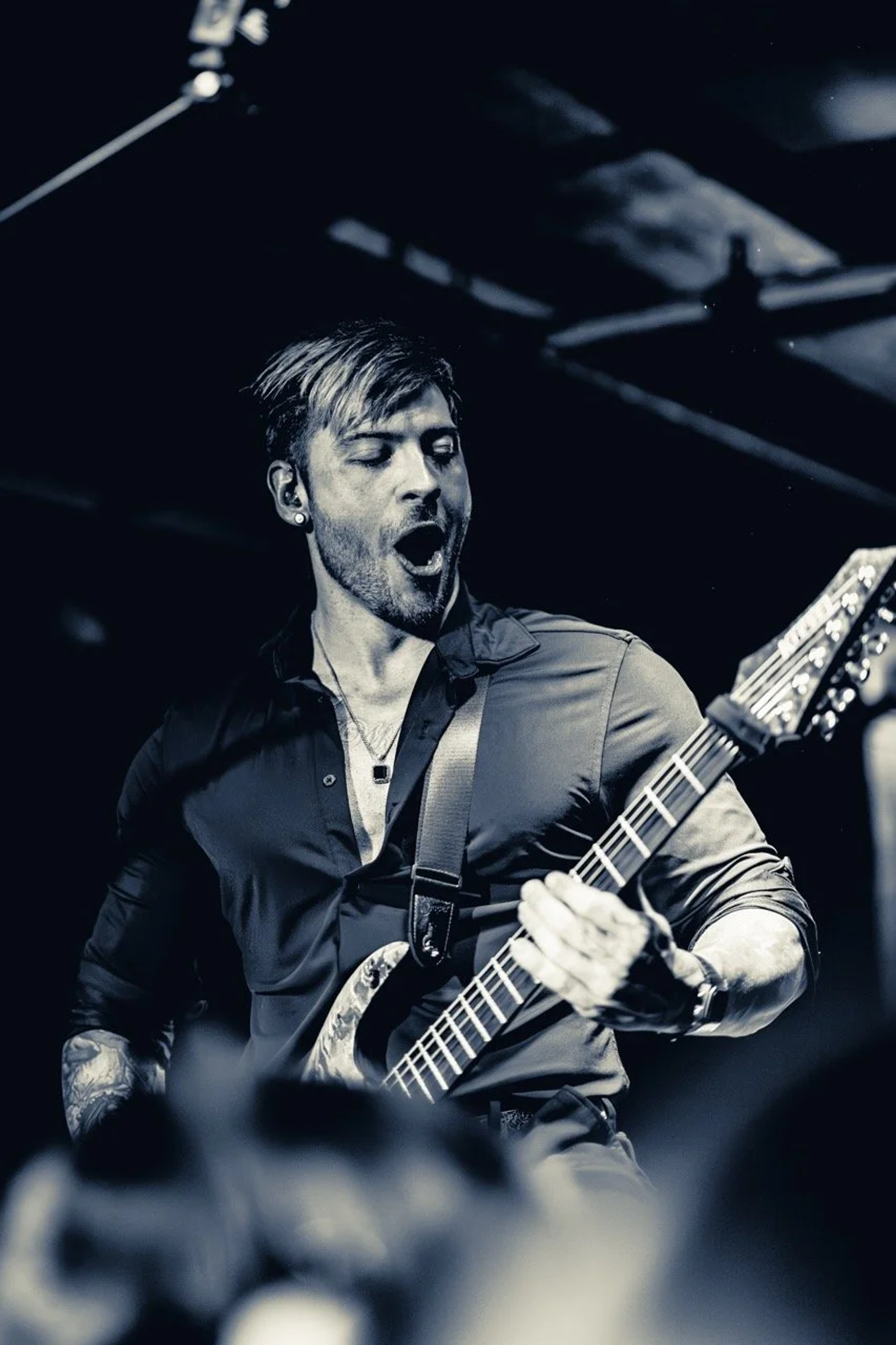 A bearded guitarist performing on stage with an electric guitar, mouth open mid-song, lit by stage lighting against a dark background.