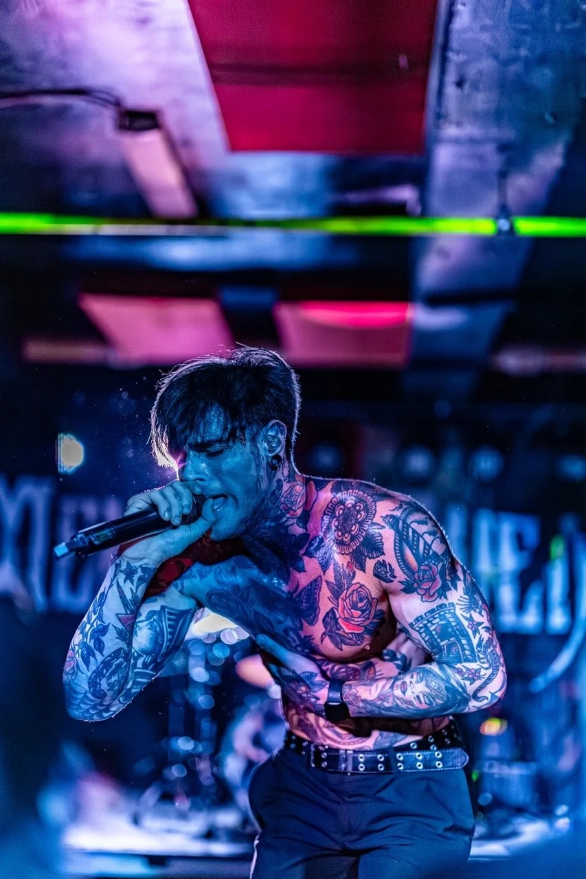 A shirtless tattooed male vocalist performs on stage holding a microphone, bathed in blue and pink stage lighting with colorful geometric displays in the background.