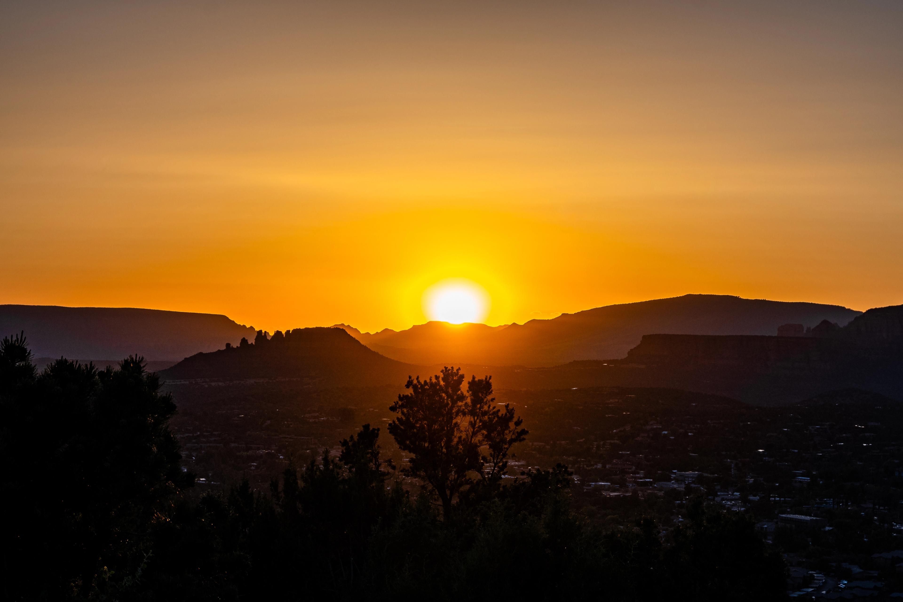 A bright sun setting between distant mesas and mountains in Sedona, Arizona, casting golden and orange light across the silhouetted landscape.