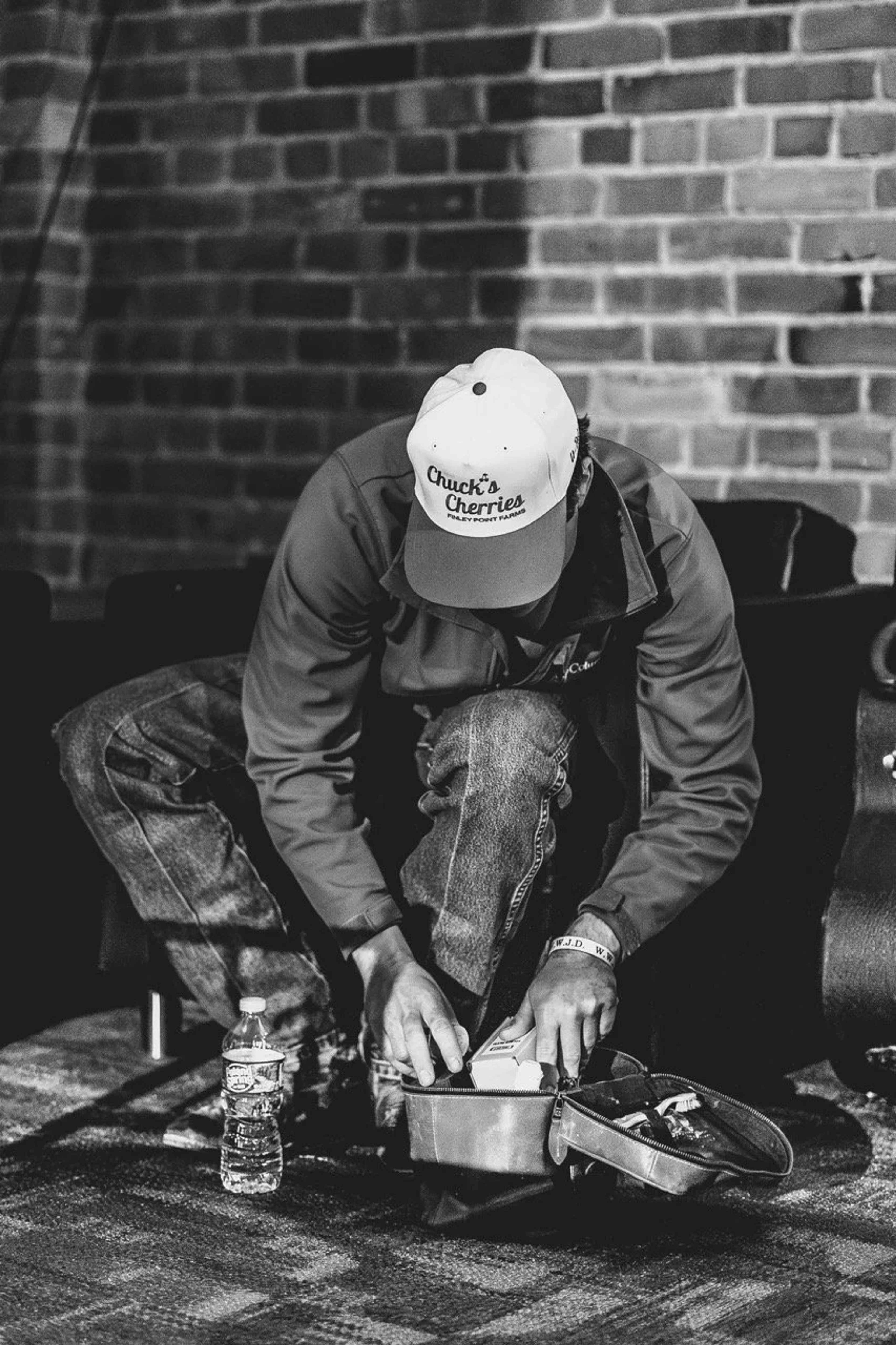A person in a Chuck's Cherries baseball cap sits crouched against a brick wall, focused on equipment on the ground, with a water bottle nearby in black and white.