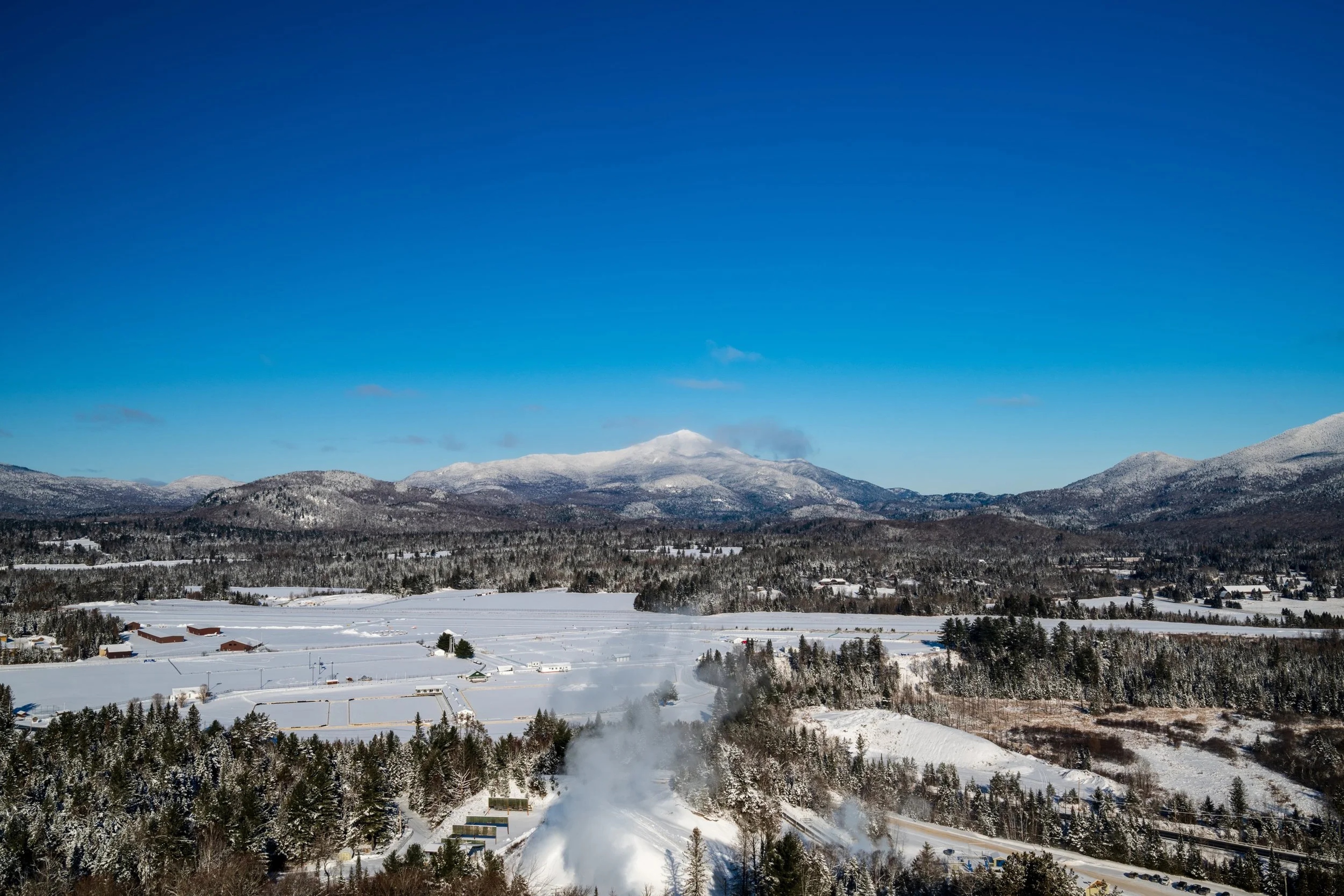 Aerial view of a snow-covered valley with a frozen lake, forested slopes, and snow-capped mountains under clear blue sky.