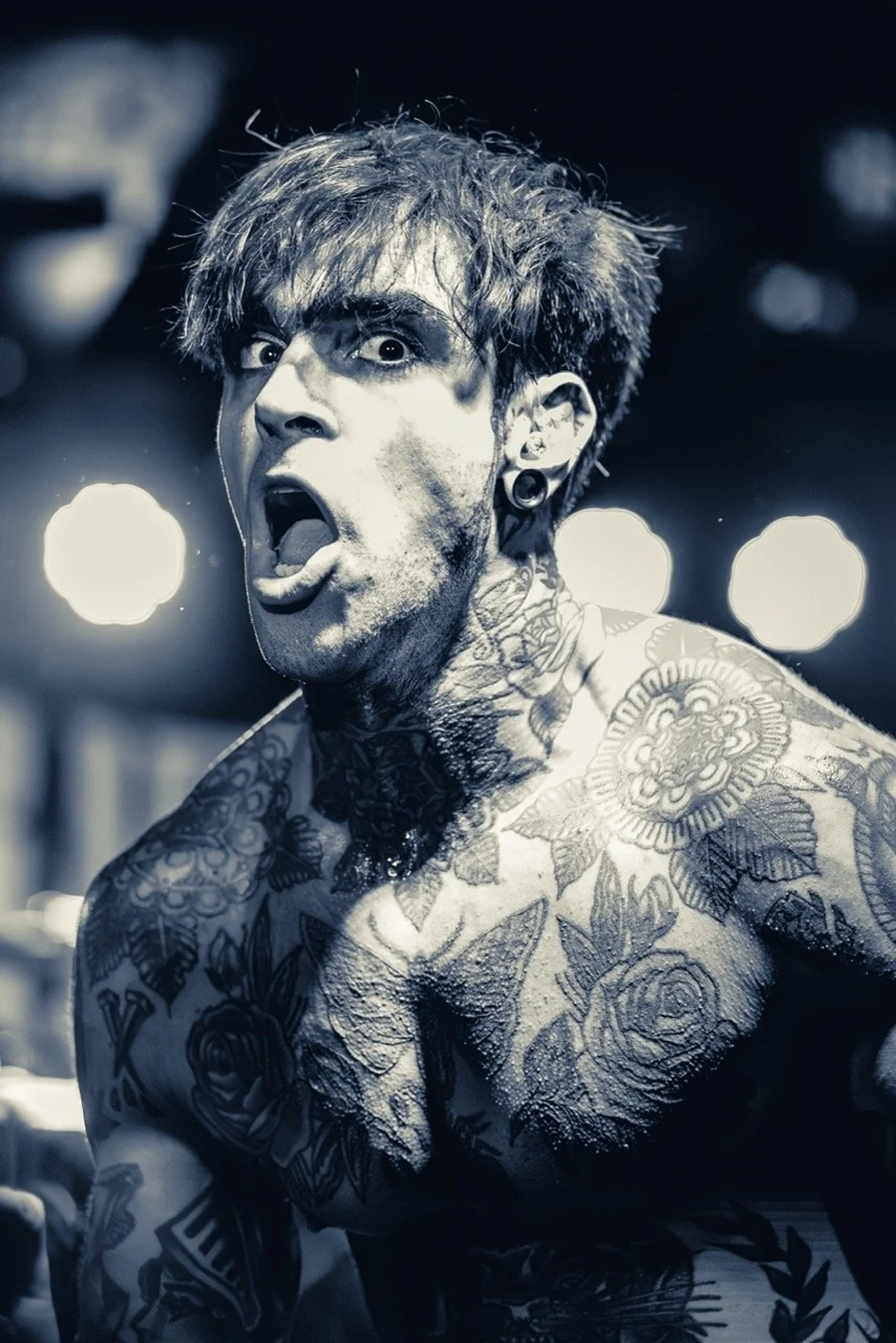 A shirtless tattooed musician on stage with mouth open mid-performance, stage lights blurred in the background, black and white photography.