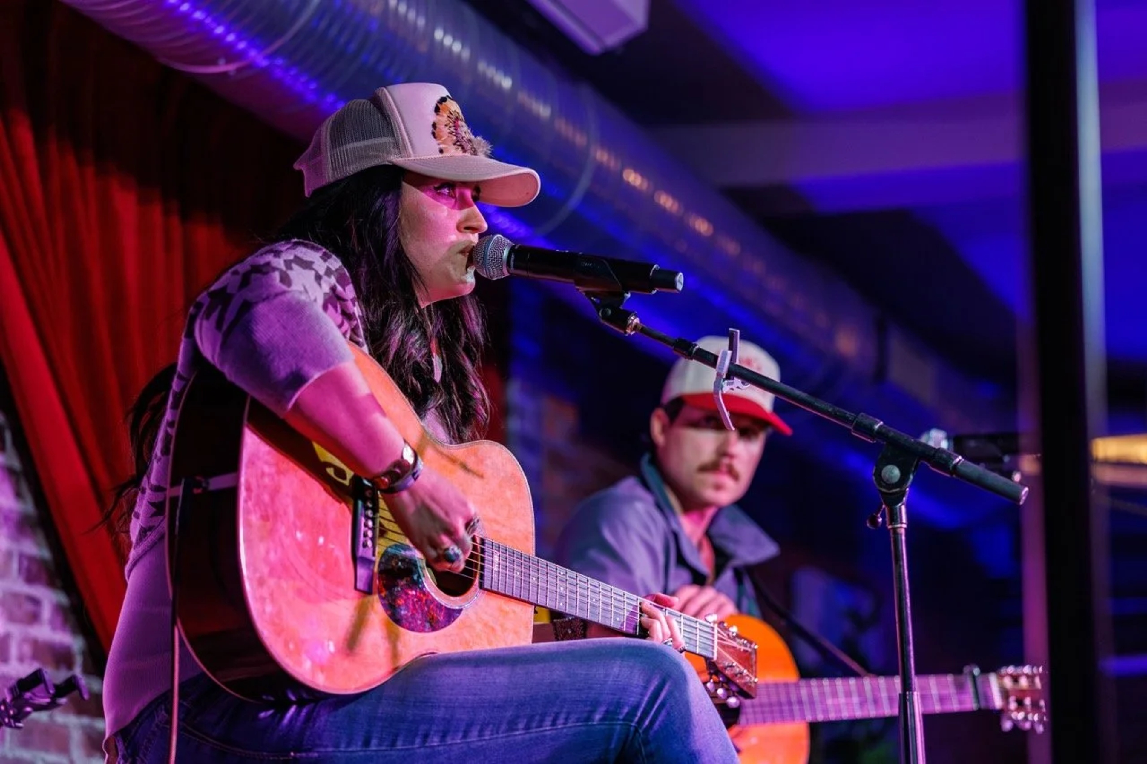 A female musician sits on stage playing an acoustic guitar and singing into a microphone, bathed in purple and pink stage lighting with a band member visible in the background.