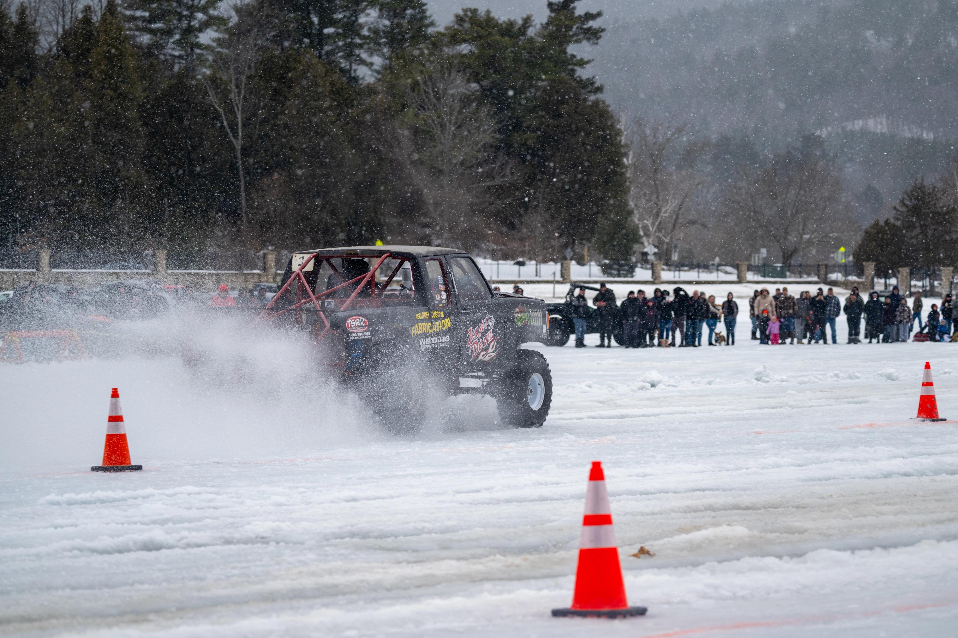 A modified truck drifting on a frozen lake, spraying snow, with spectators watching from the shoreline behind safety cones.