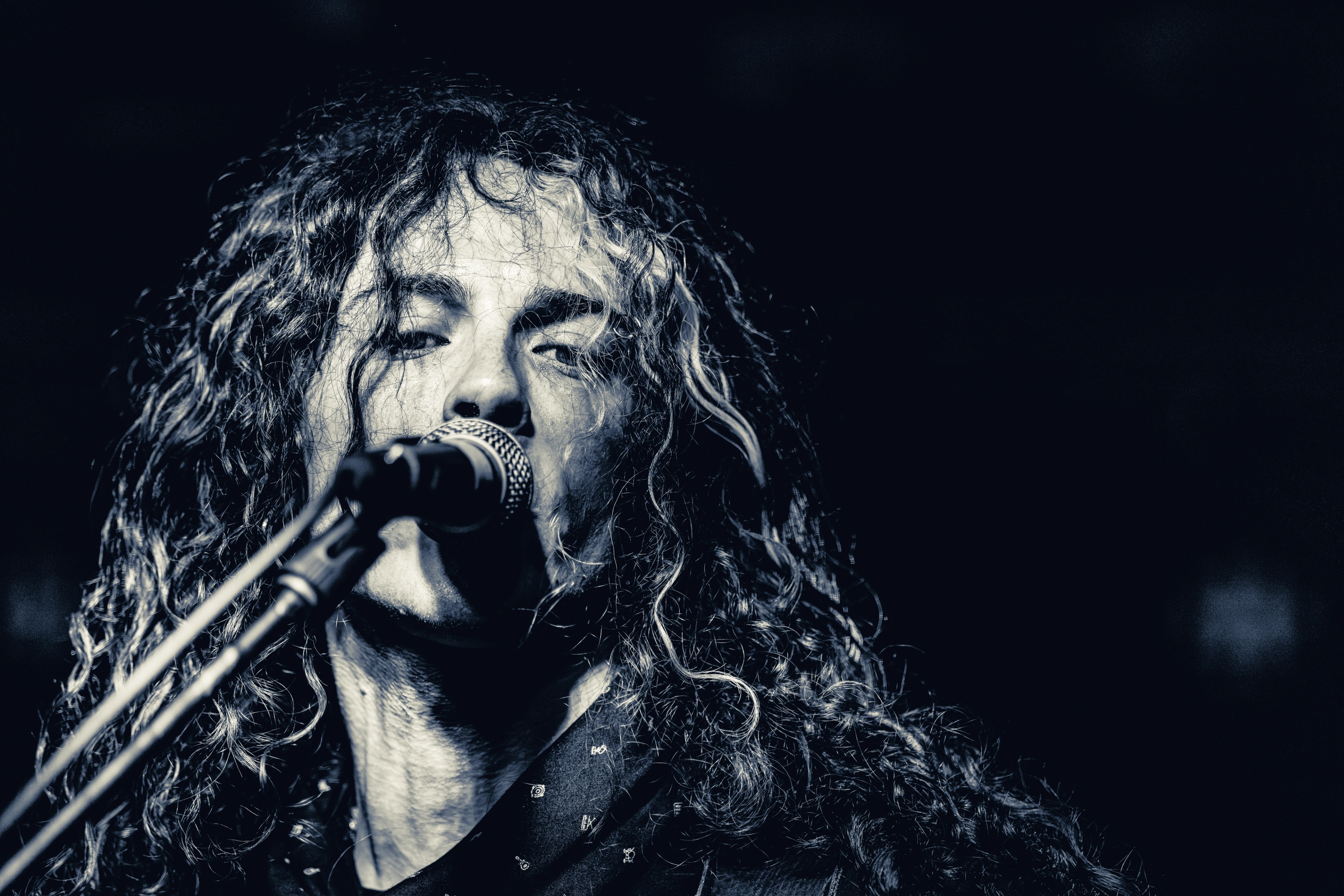 A long-haired singer performing at a microphone in monochromatic lighting, eyes closed, fully committed to the moment.