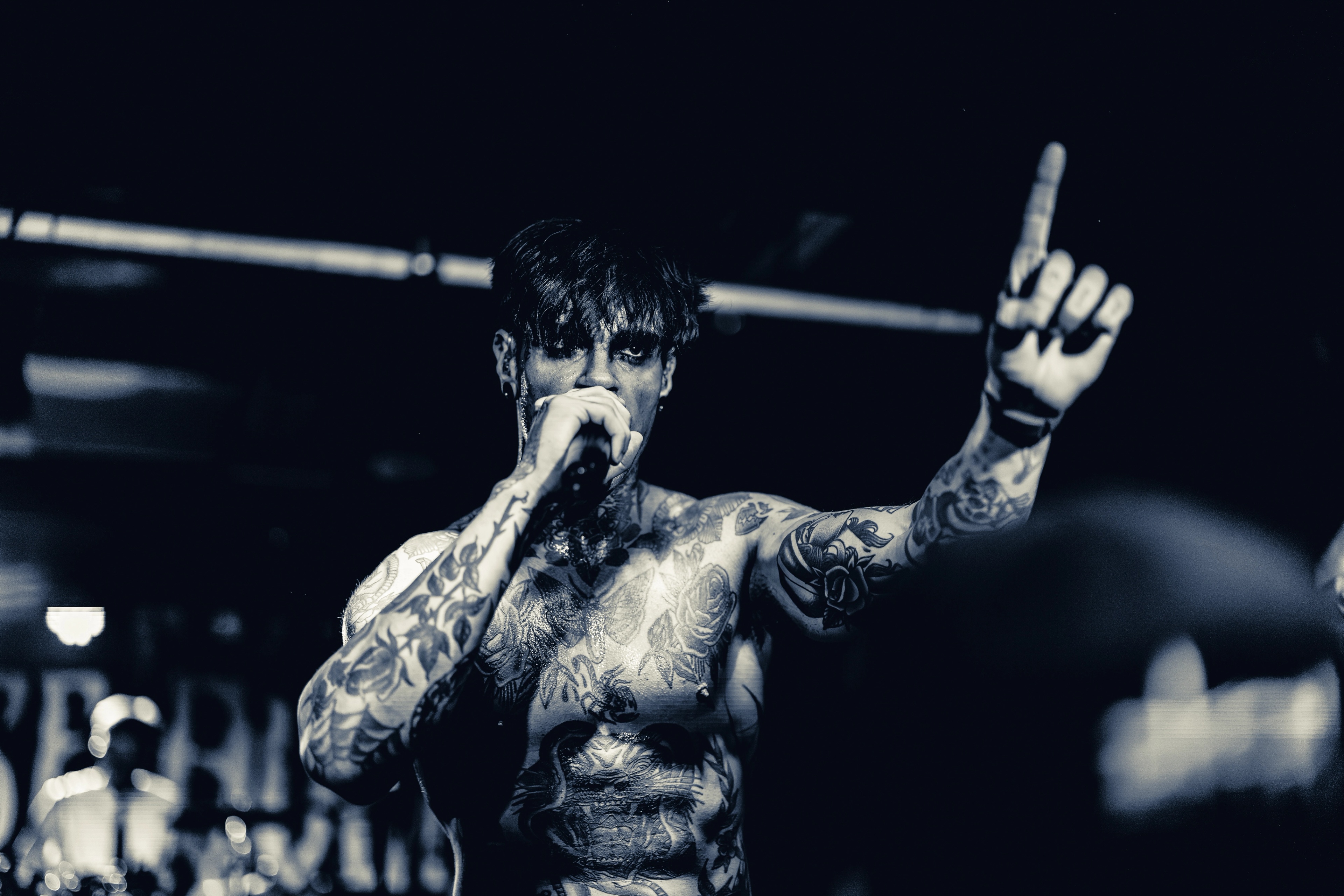 A shirtless tattooed musician on stage holding a microphone, arm raised, performing under stage lighting at an indoor venue.