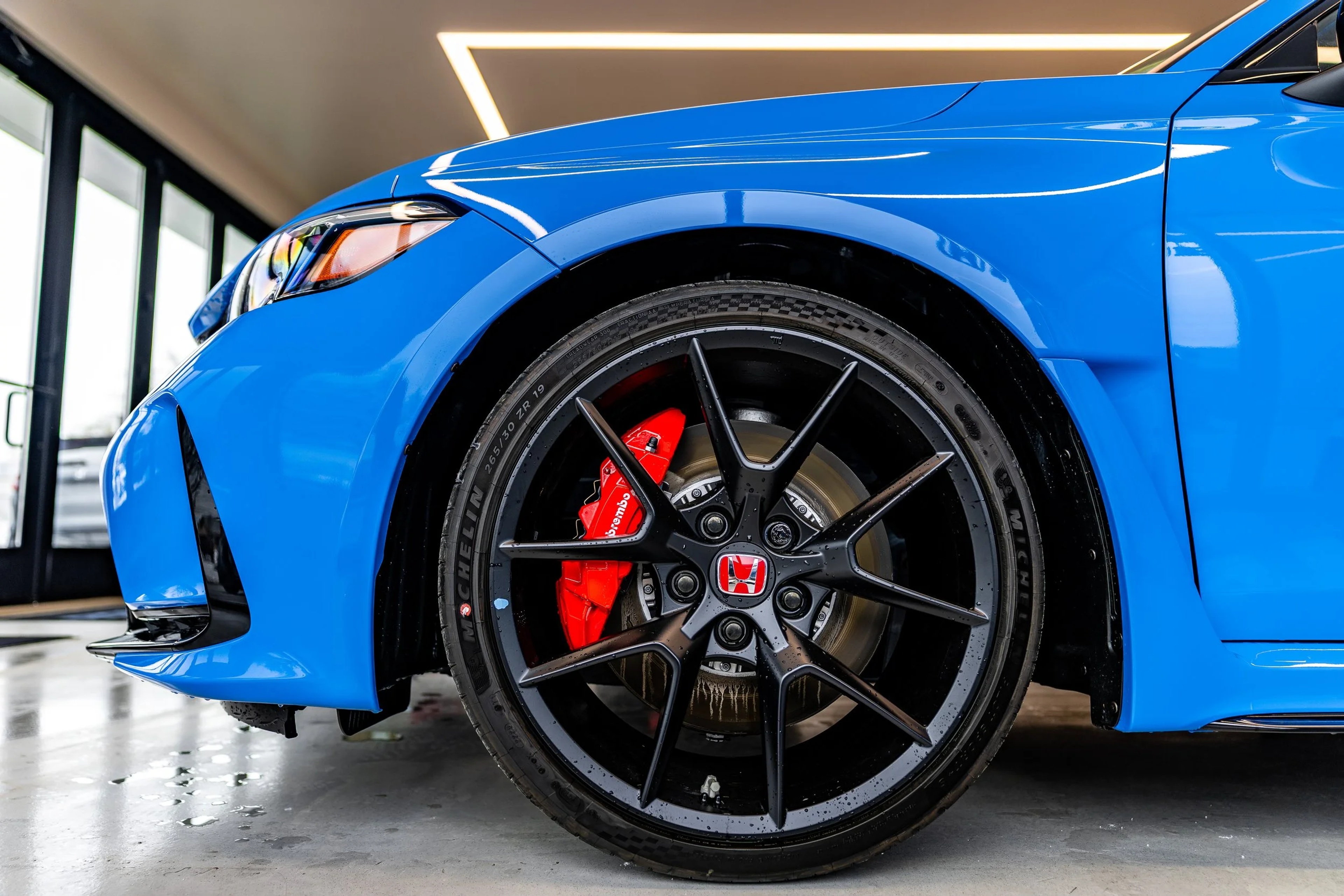 Close-up of a bright blue Honda sedan showing a black multi-spoke wheel with red brake calipers, photographed in an indoor showroom with white lighting.
