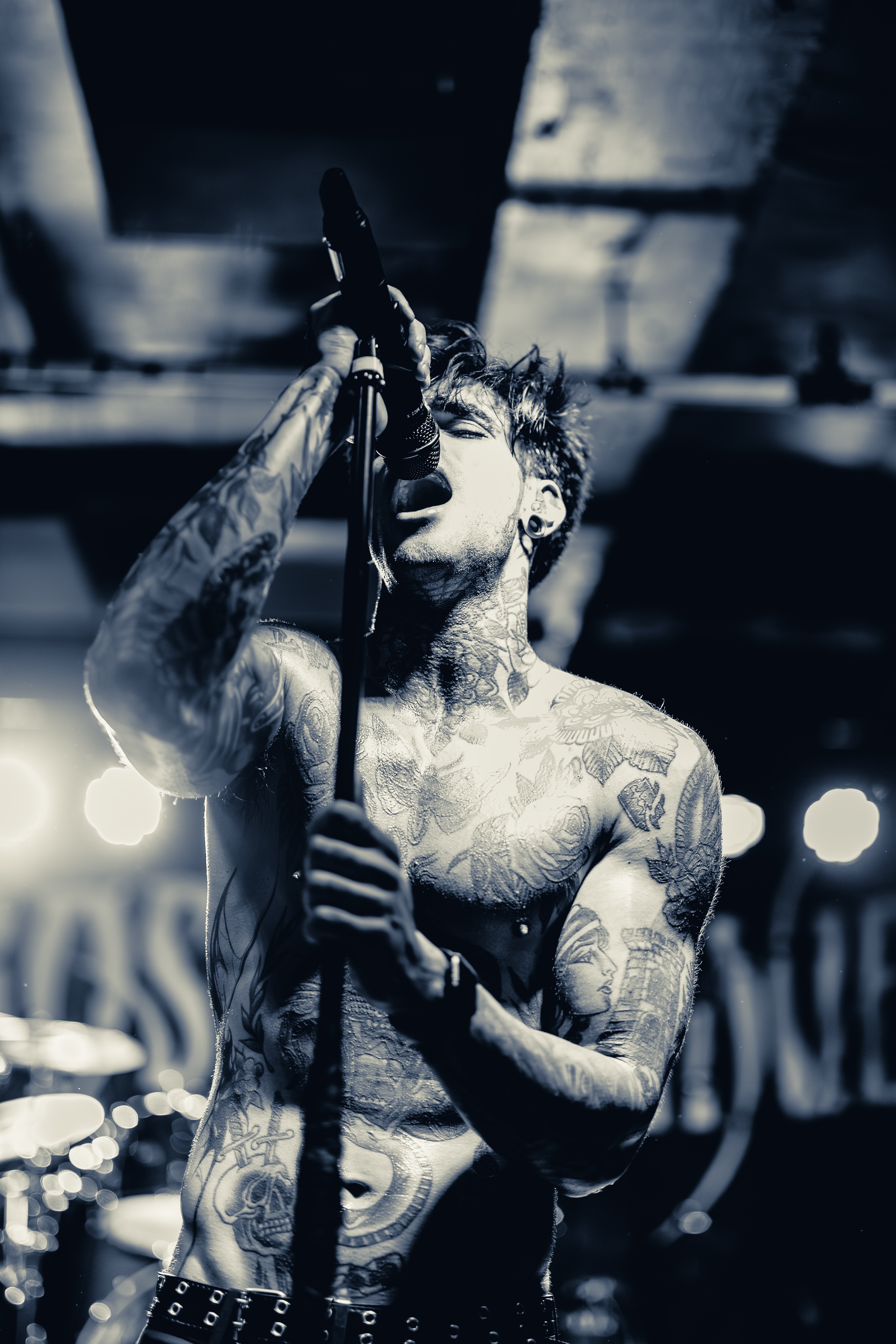A shirtless tattooed vocalist performs at a microphone on stage, mouth open mid-song, arm raised, with blurred stage lights and industrial backdrop.