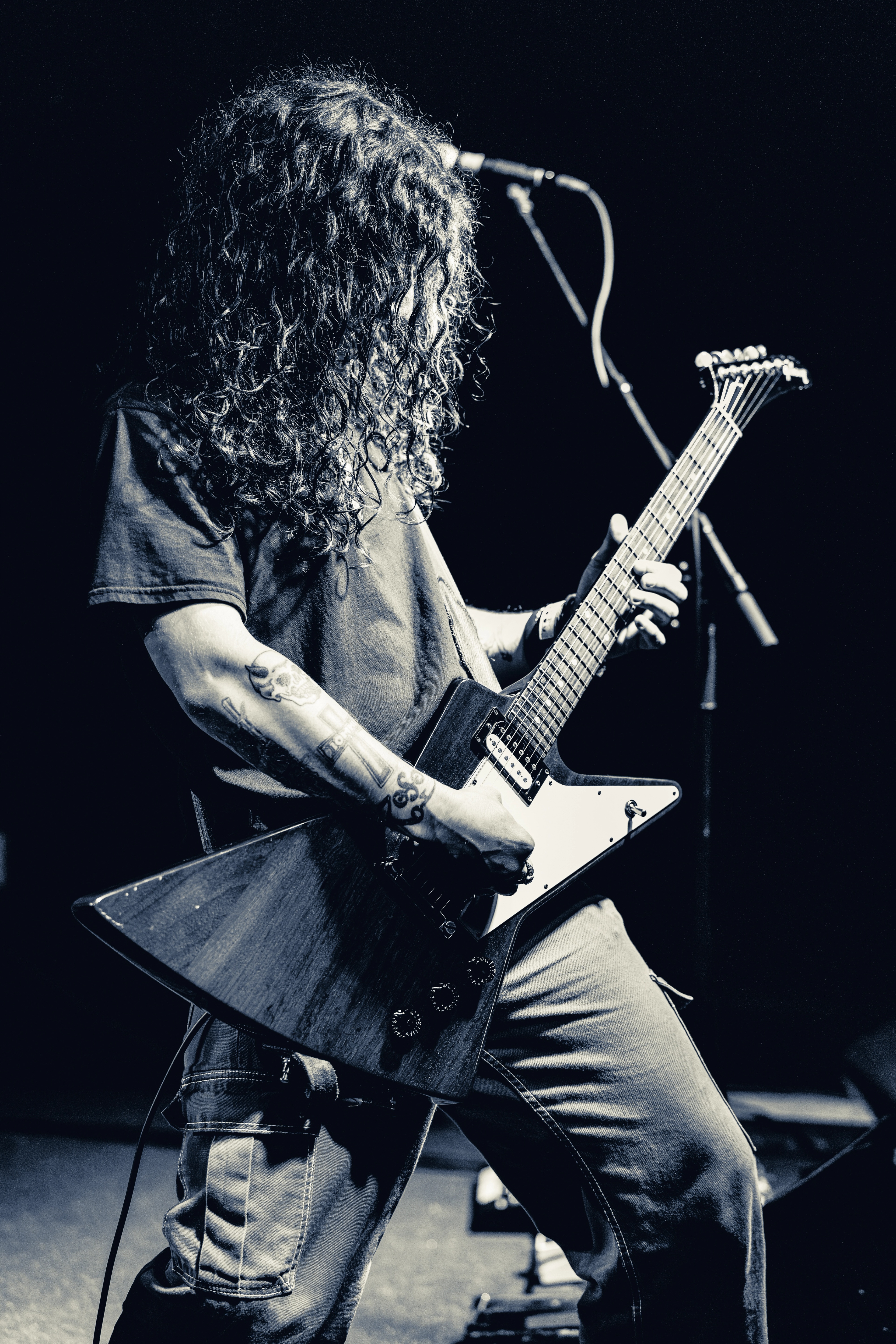 A long-haired guitarist in a denim shirt plays a white electric guitar on stage, head down, illuminated against a dark background with a microphone visible.