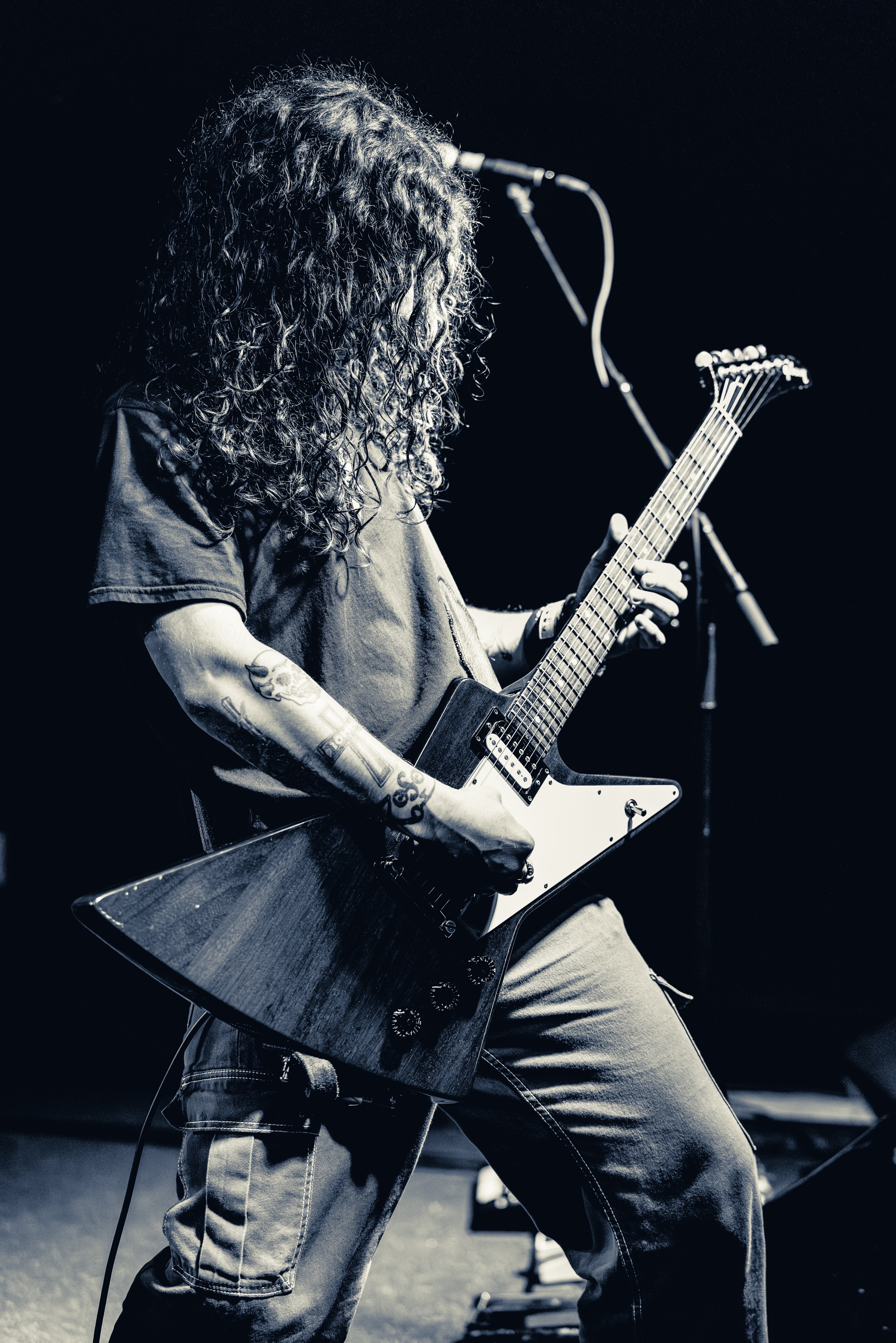 A long-haired guitarist in a denim shirt plays a white electric guitar on stage, head down, illuminated against a dark background with a microphone visible.