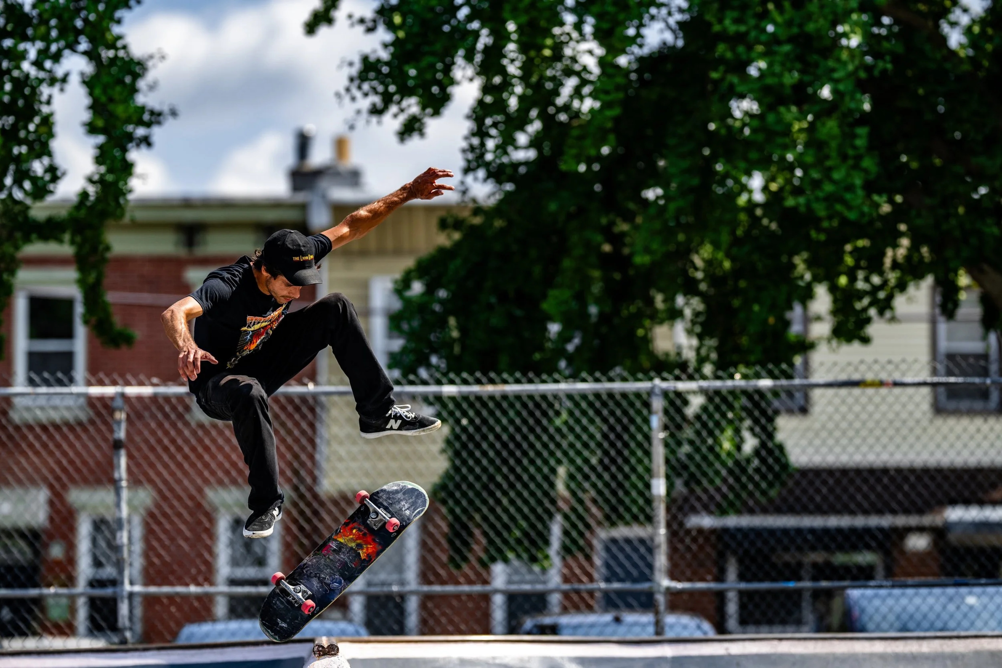 A skateboarder performs an aerial trick on a concrete court, captured mid-flight with arm extended, surrounded by chain-link fencing and ivy-covered buildings.
