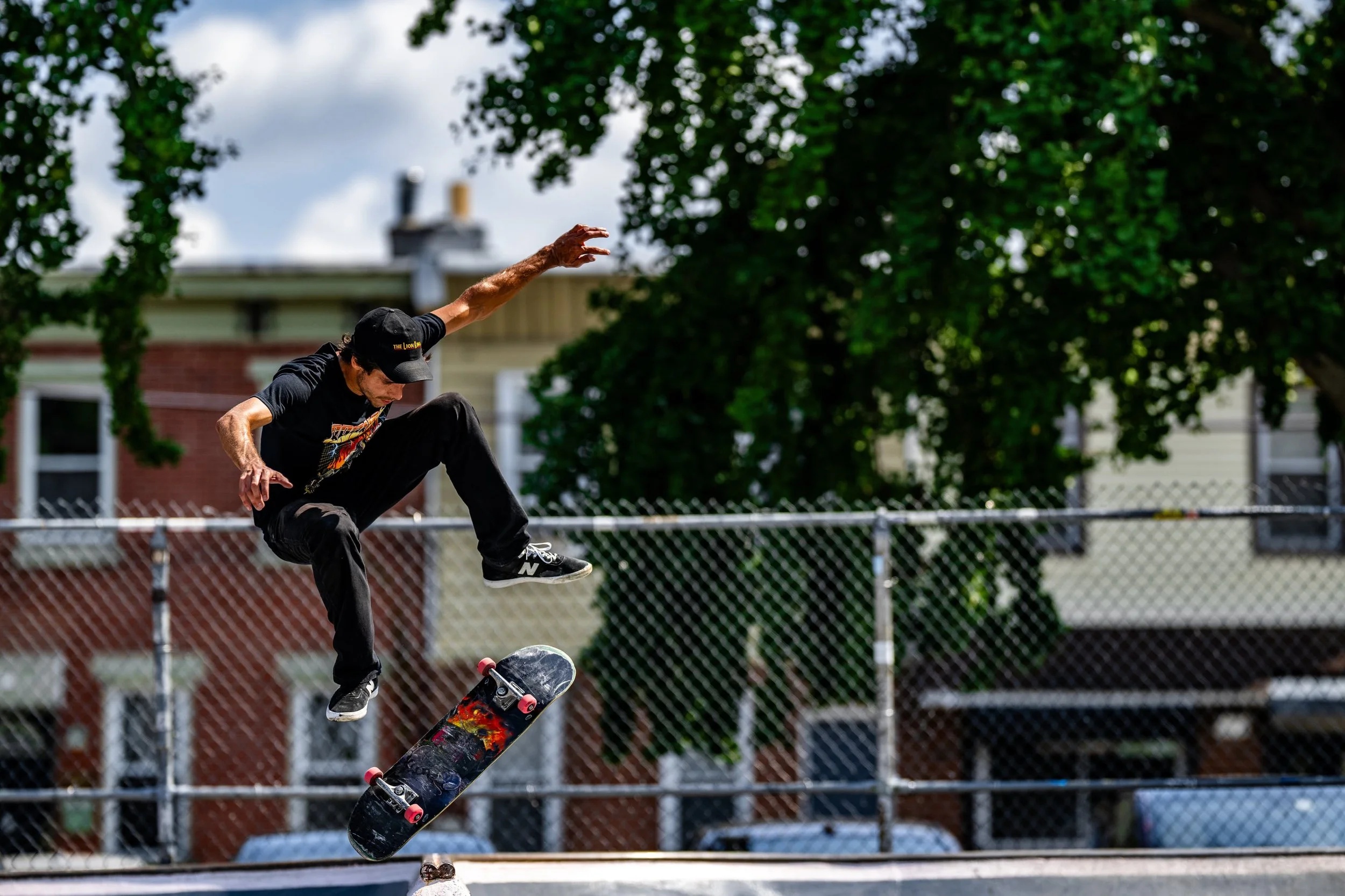 A skateboarder performs an aerial trick on a concrete court, captured mid-flight with arm extended, surrounded by chain-link fencing and ivy-covered buildings.