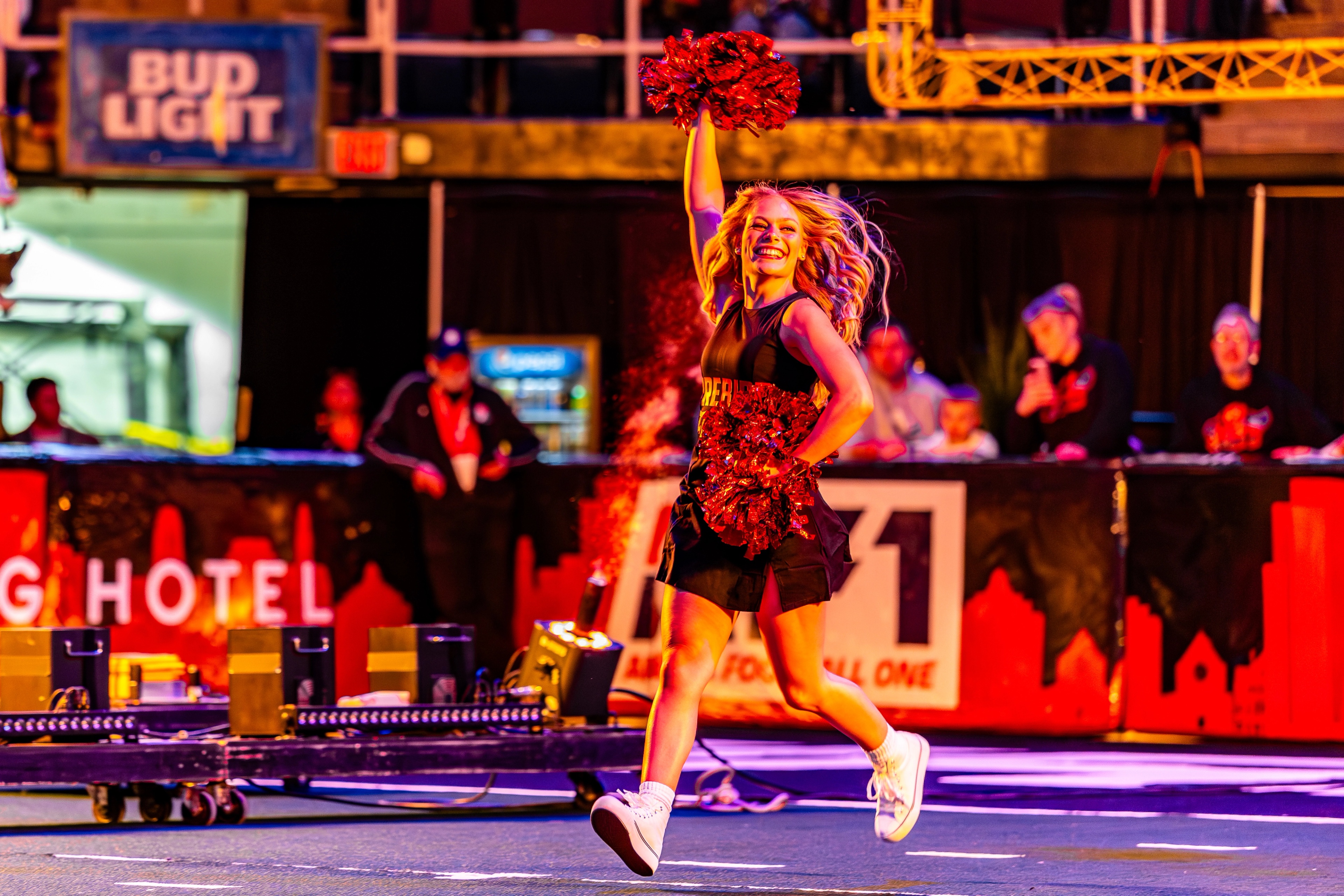 A cheerleader in a red sequined uniform leaps across an arena floor, pom-poms raised, with stadium lighting and signage visible in the background.
