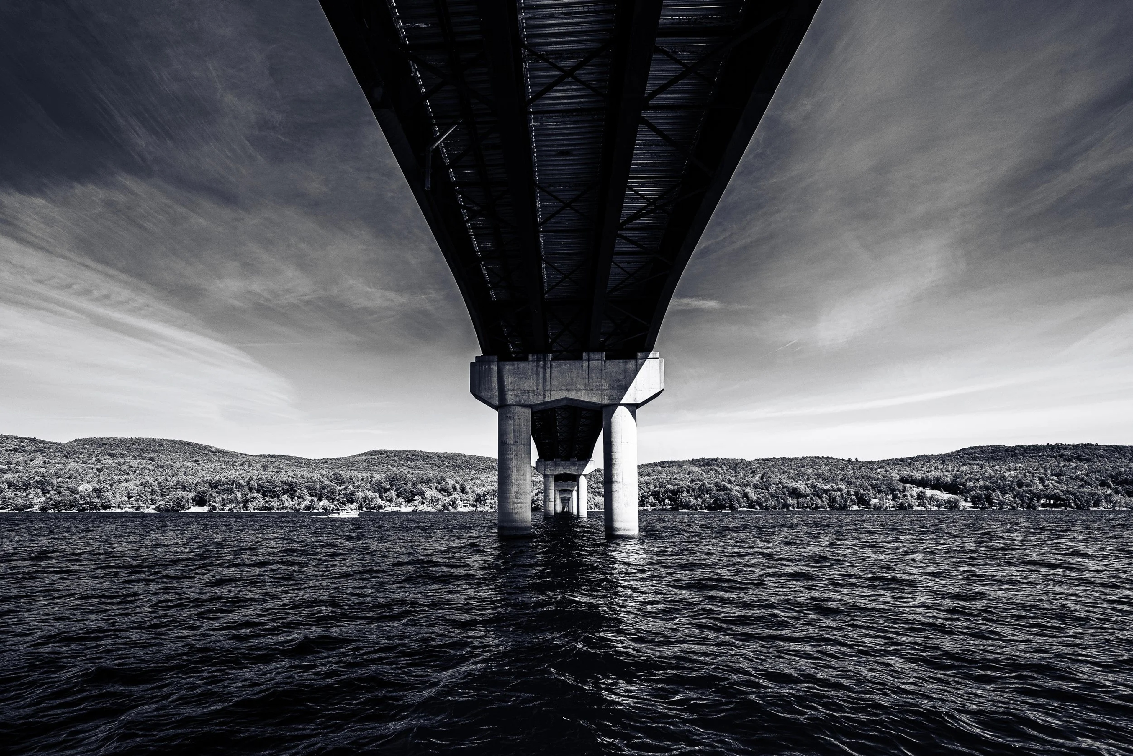 A bridge spans across a lake with forested hills in the background, shot from directly underneath the structure in black and white.