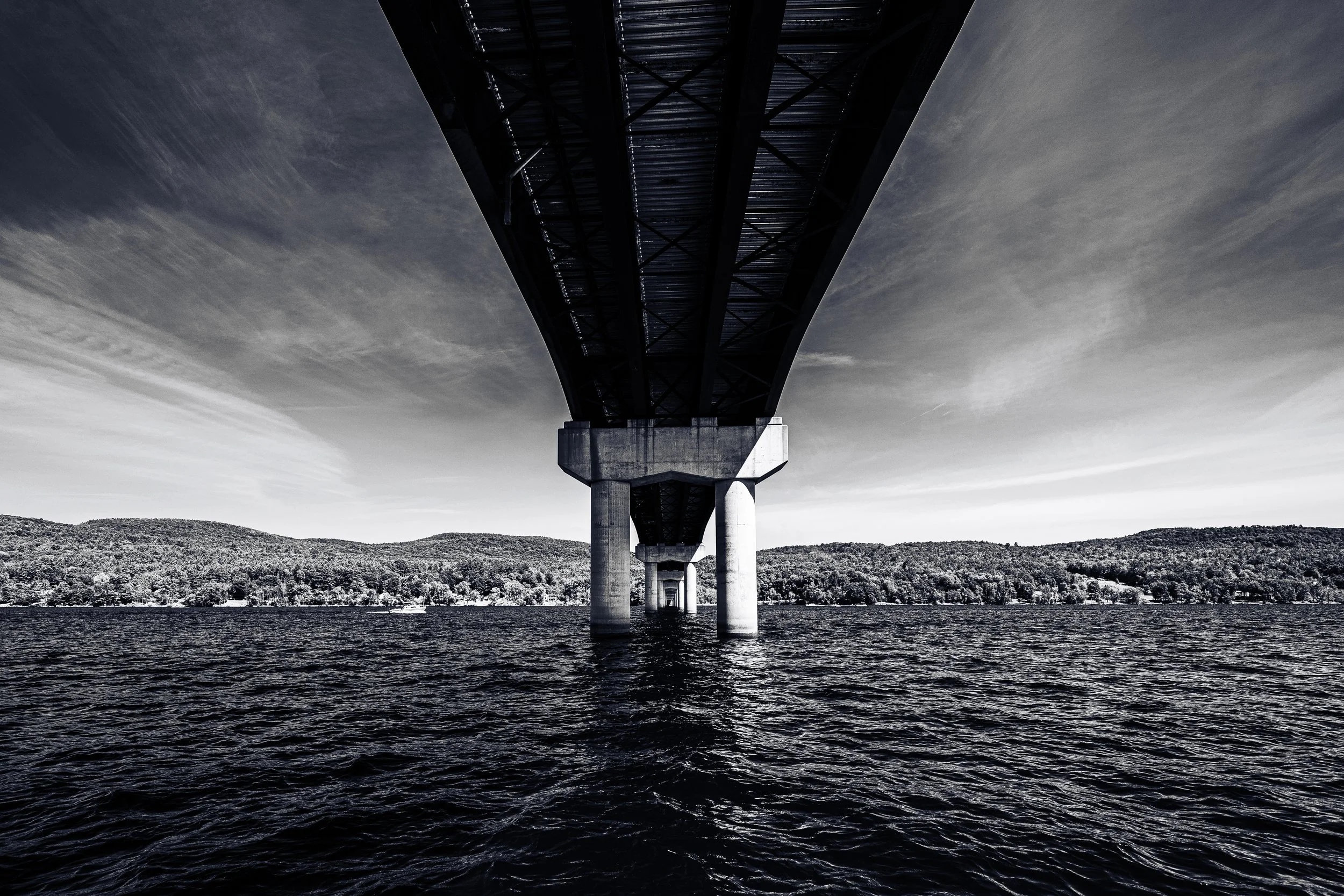 A bridge spans across a lake with forested hills in the background, shot from directly underneath the structure in black and white.