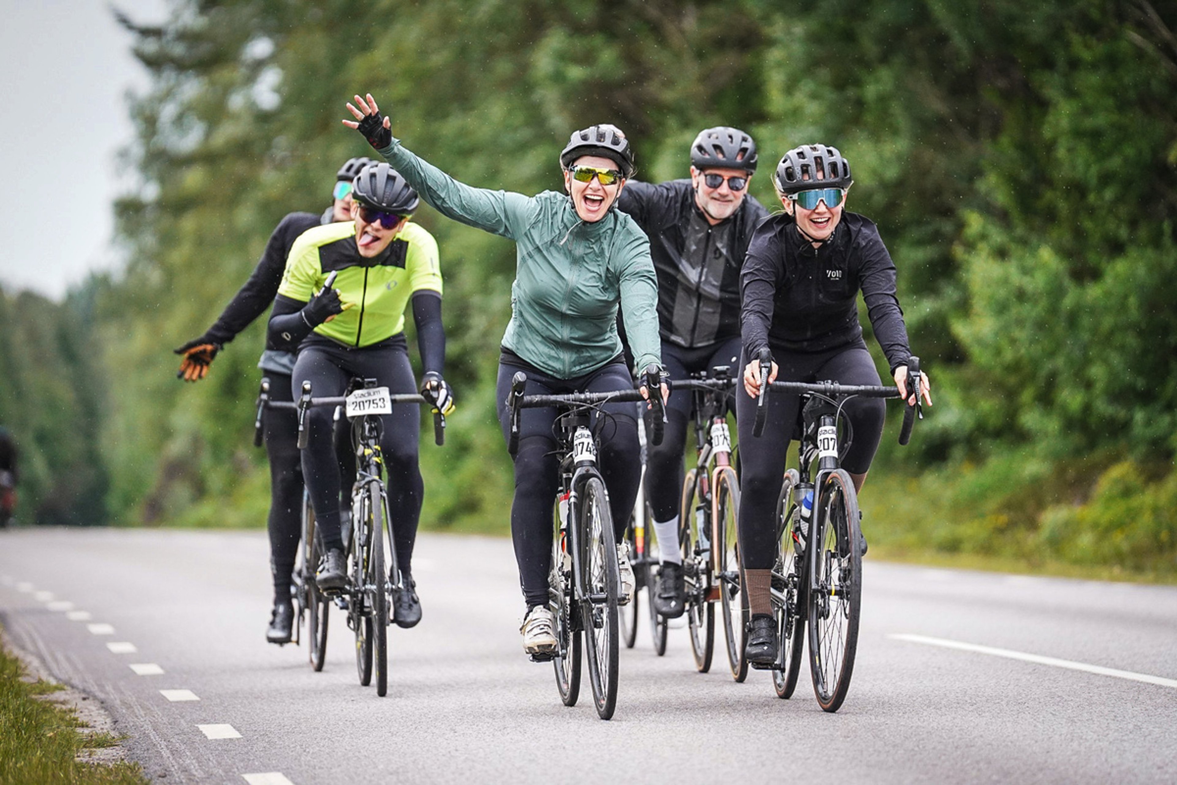 A group of five cyclists, with two women in the foreground smiling and waving, ride down a tree-lined road.