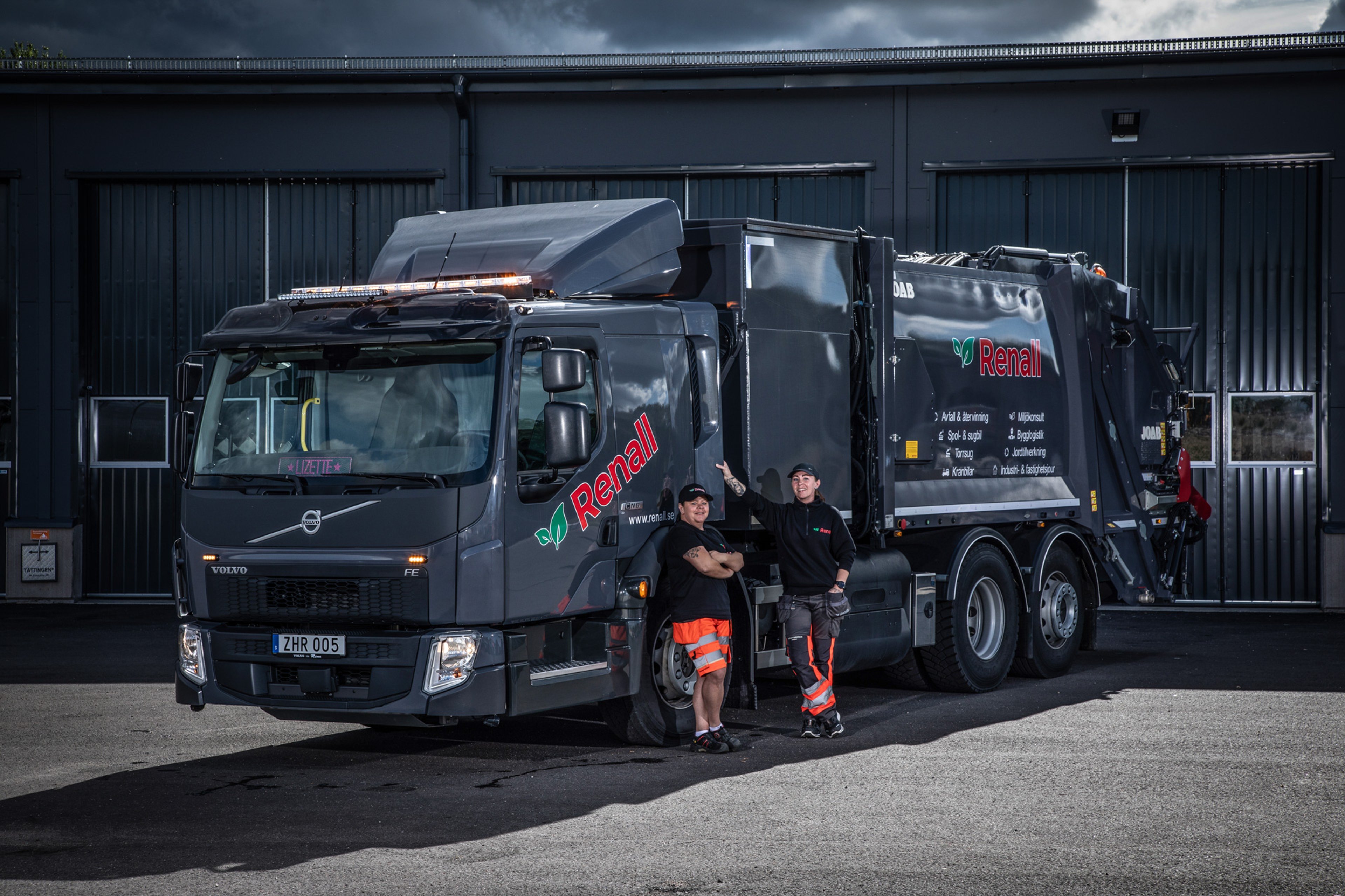 Two persones in work attire stand next to a dark grey Renall garbage truck in front of a building.