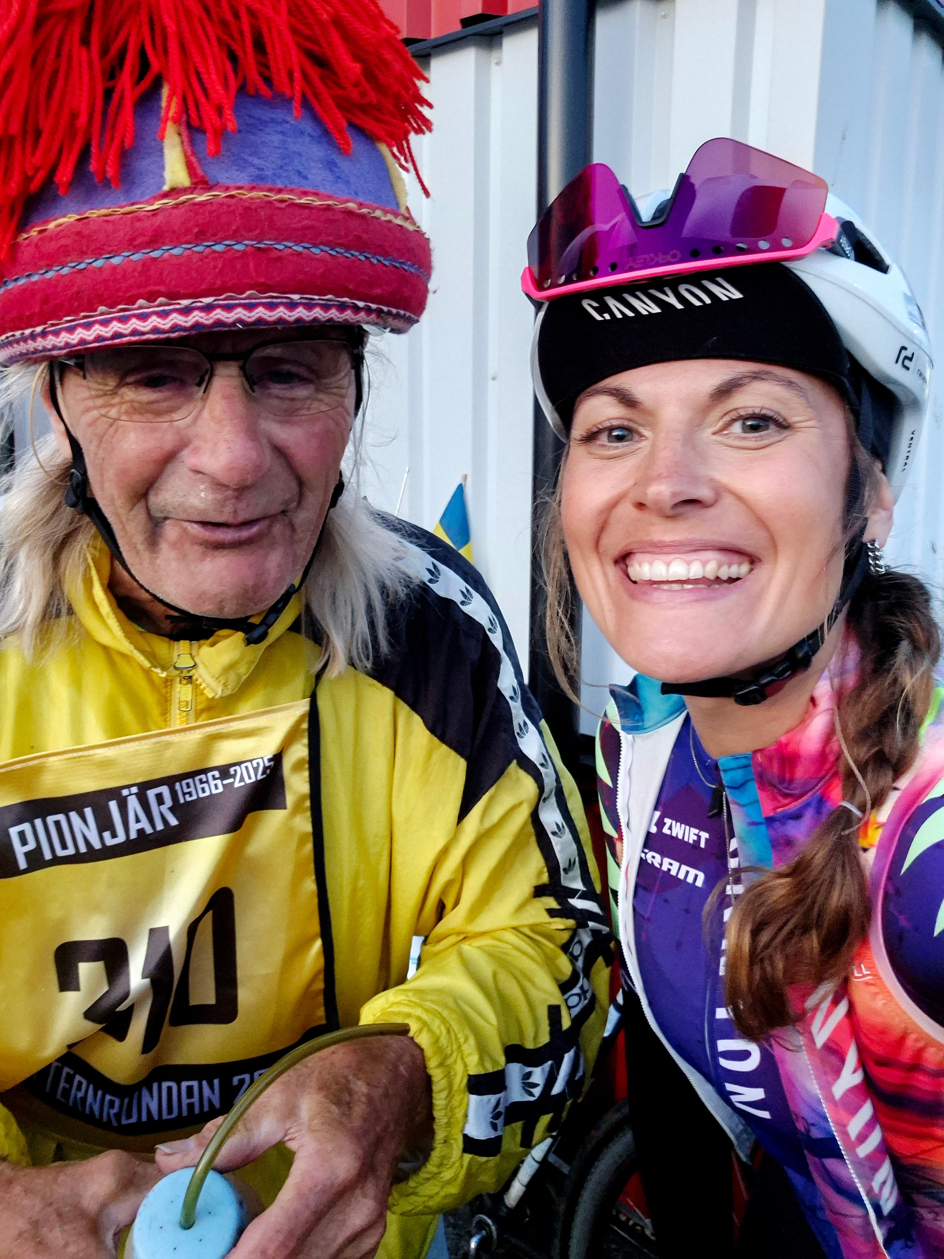 Two smiling cyclists, an older man in a traditional colorful hat with red tassels and a yellow jacket, and a woman in a helmet and colorful jersey.