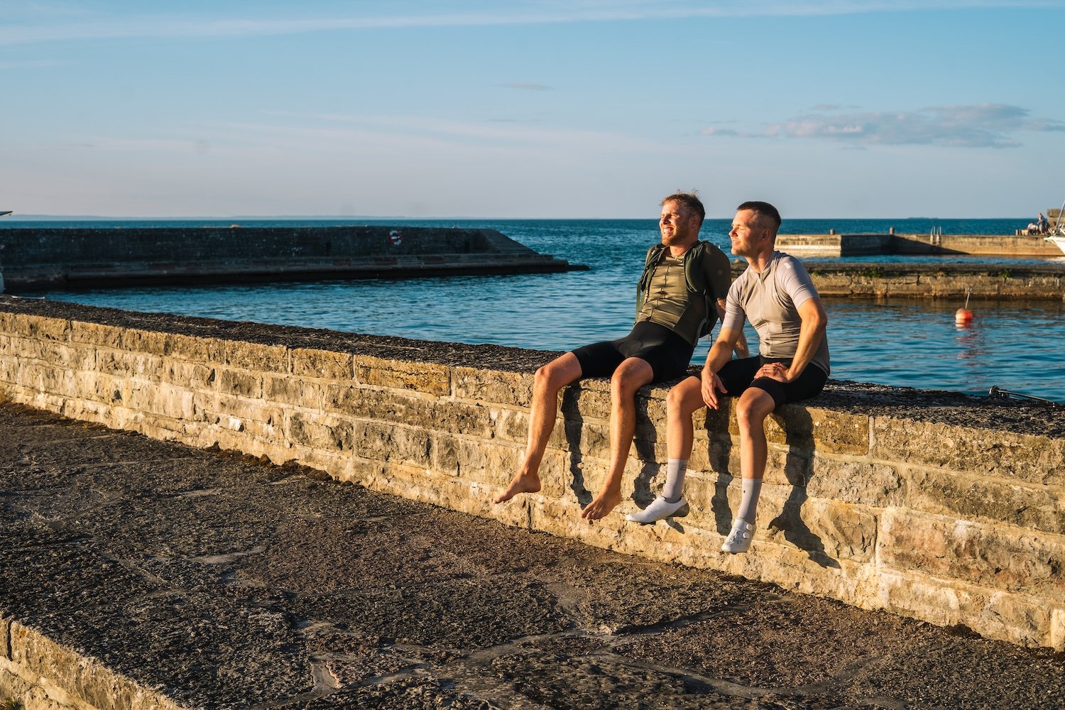 Two men sit on a stone wall overlooking water, one with a prosthetic arm.