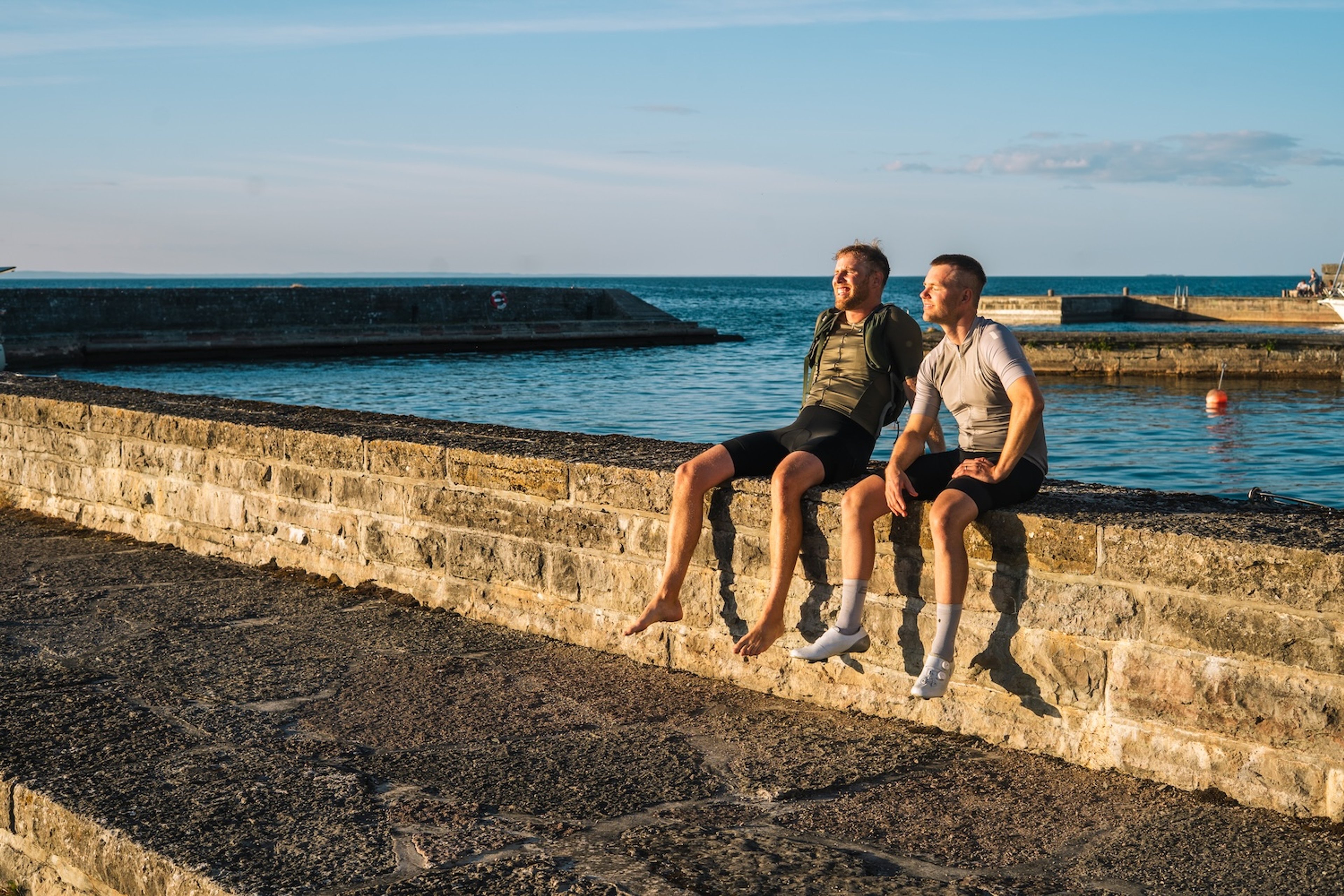 Two men sit on a stone wall overlooking water, one with a prosthetic arm.