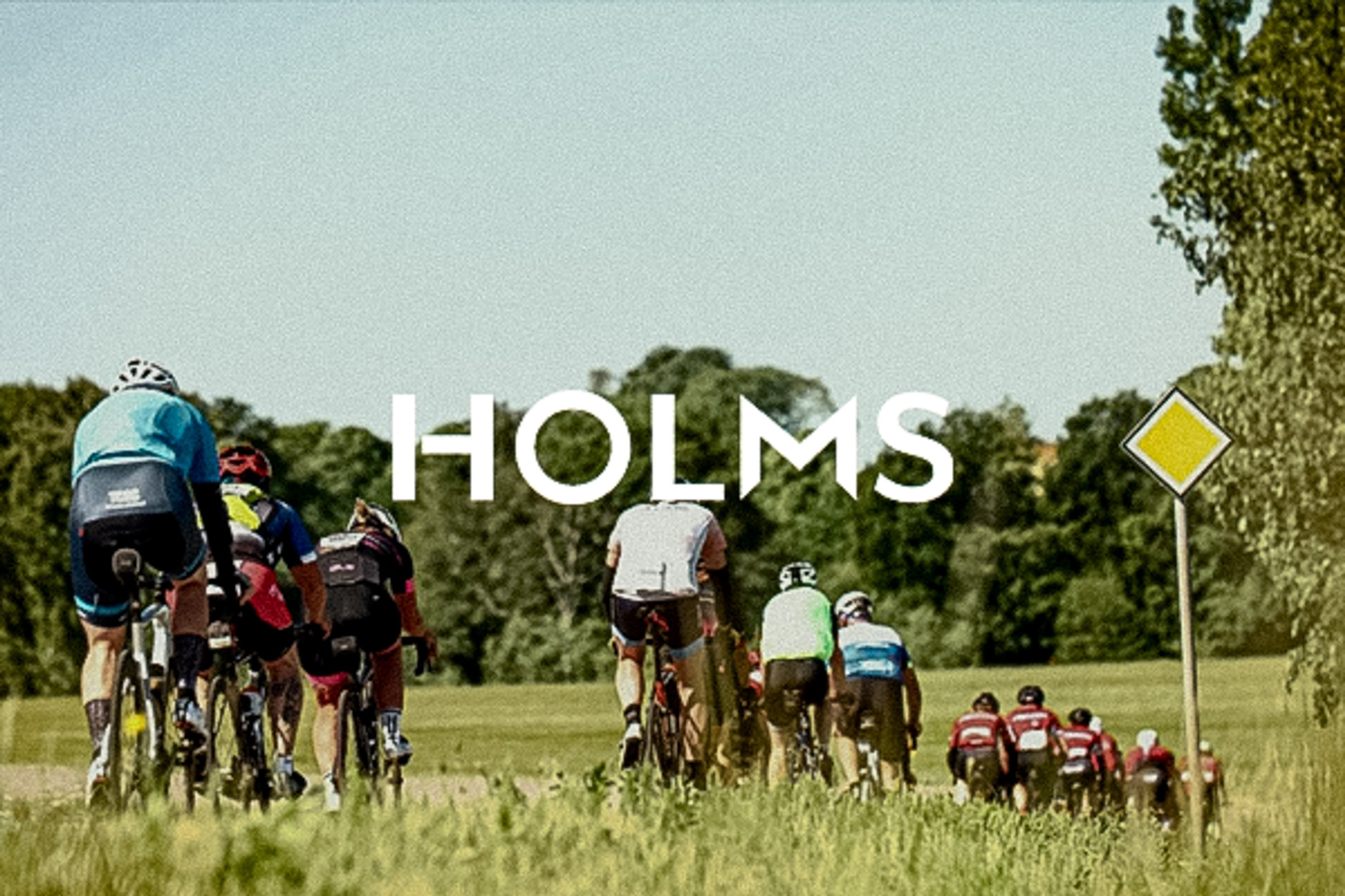 Group of cyclists riding away on a rural road with trees and a road sign, with "HOLMS" overlaid.