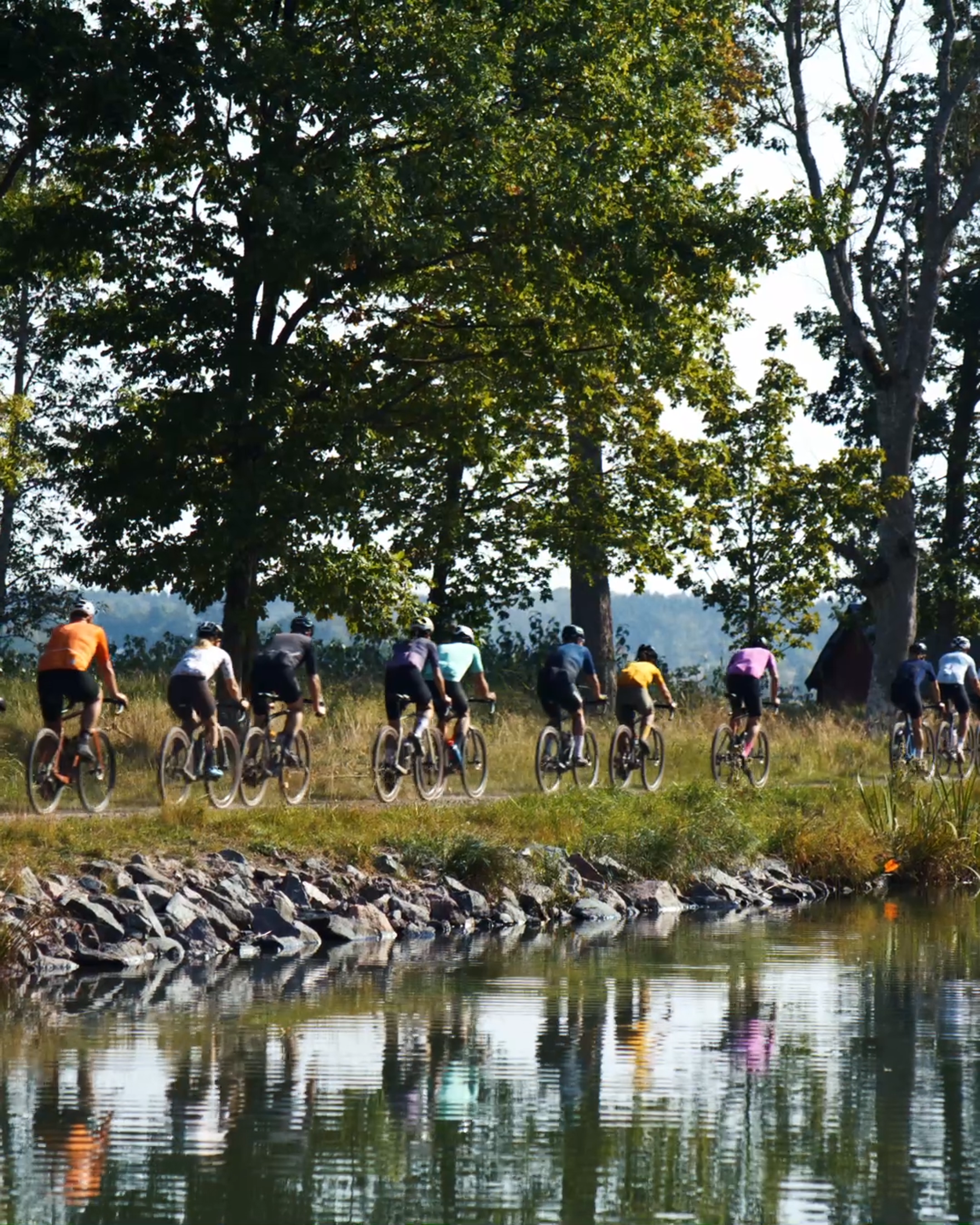 A line of cyclists rides along a dirt path beside a canal, reflecting the cyclists and trees.