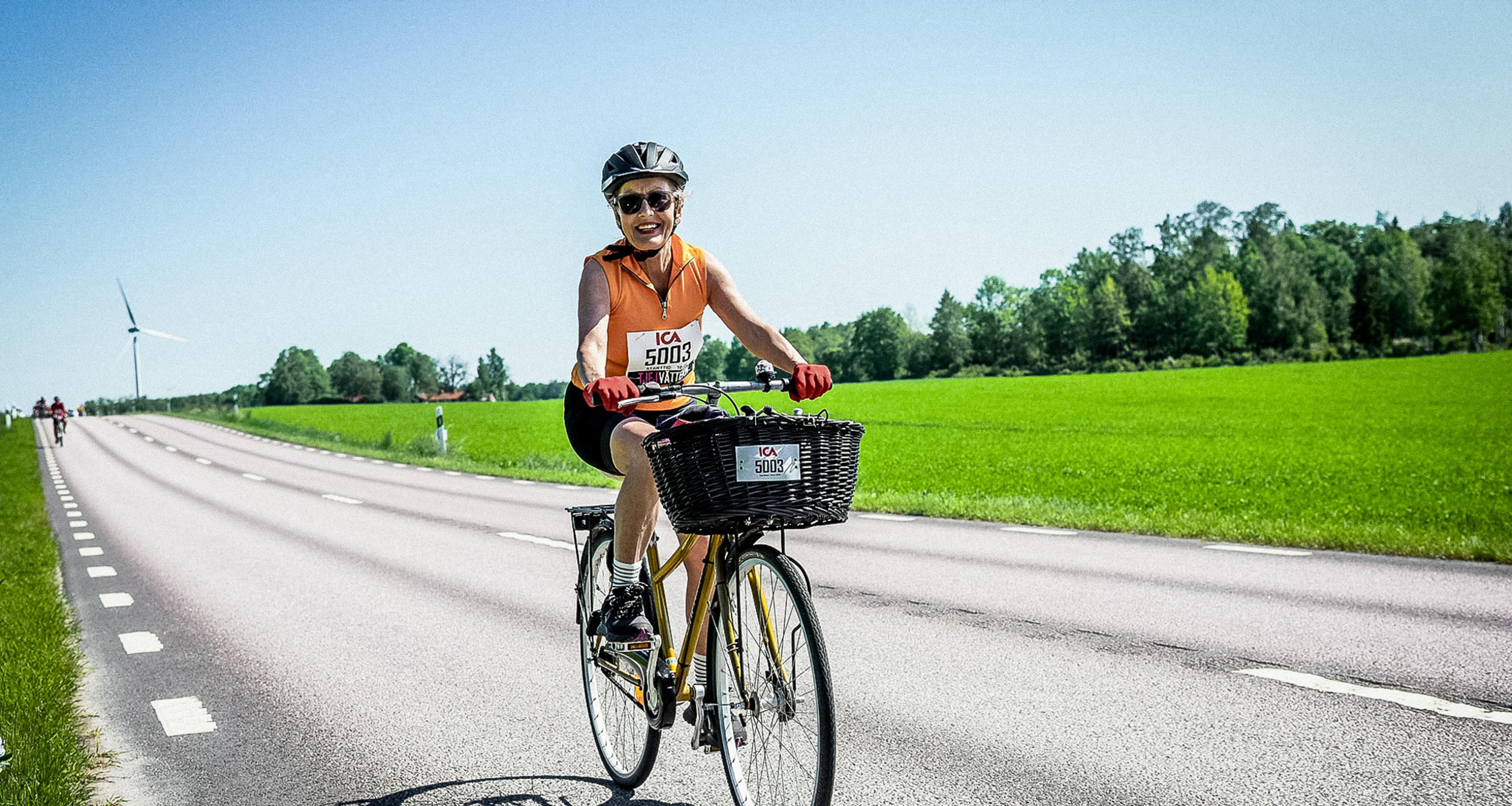 A woman in a helmet and sunglasses smiles while riding a bicycle with a front basket on a sunny road, with green fields and a wind turbine in the background.