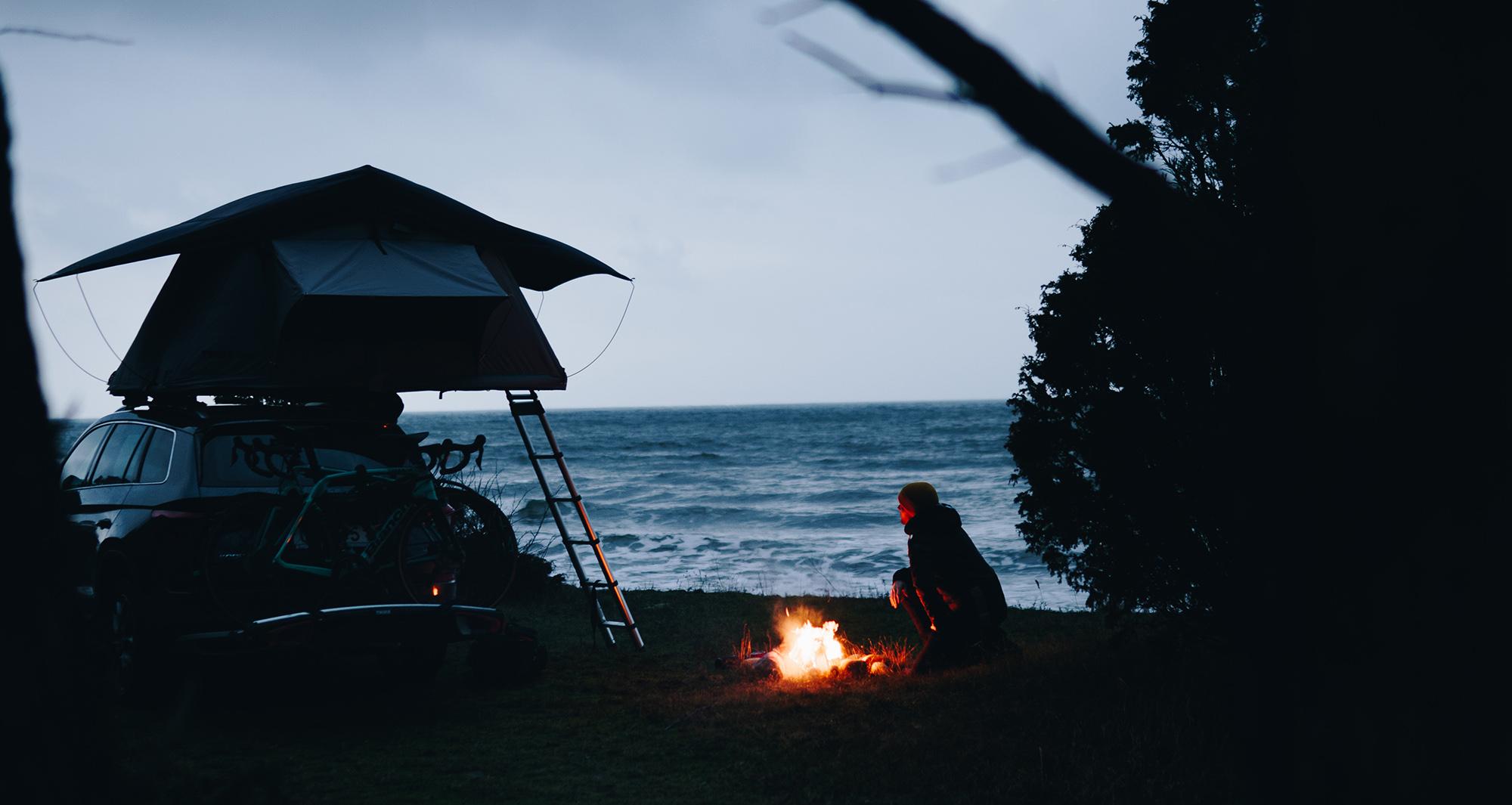 A person sits by a campfire next to a car with a rooftop tent and bikes, overlooking the ocean at dusk.
